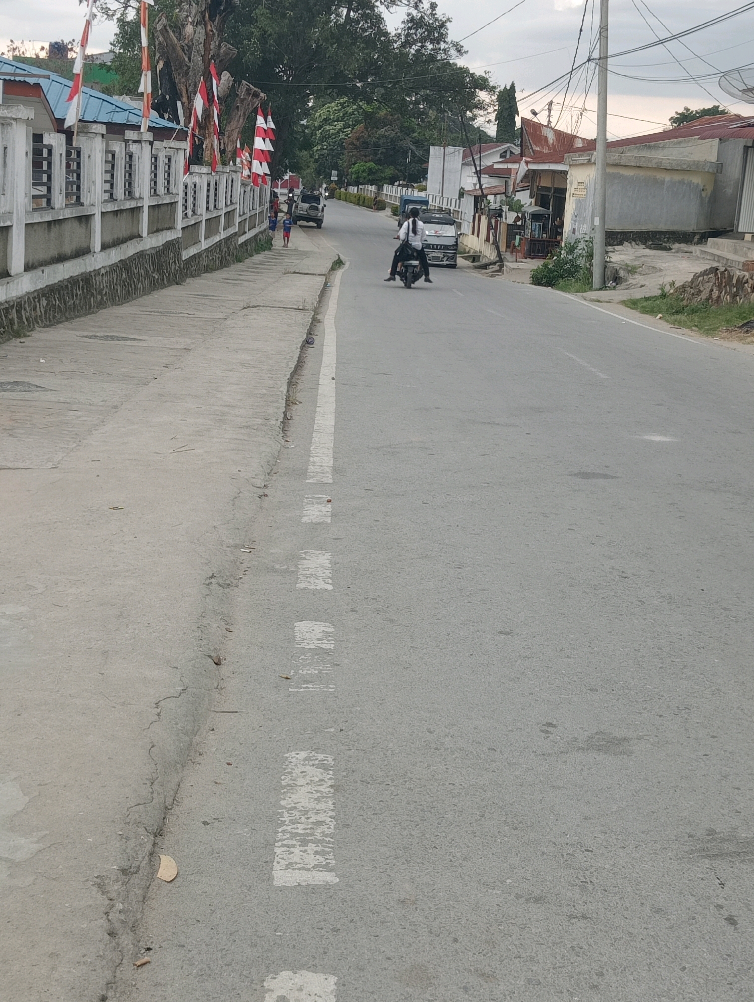 The image depicts a street scene in Hinalangbagasan, Indonesia. A motorcycle is being ridden by a person wearing a white shirt and dark pants, with a passenger also in a white shirt. The motorcycle is on the right side of the road. On the left side of the road, a concrete sidewalk runs alongside a low wall decorated with Indonesian flags. Two children, one in a blue shirt and the other in a red shirt, are walking on the sidewalk, and a white SUV is parked behind them. Buildings with tin roofs are visible in the background, along with trees and utility poles with overhead wires. The sky is overcast.