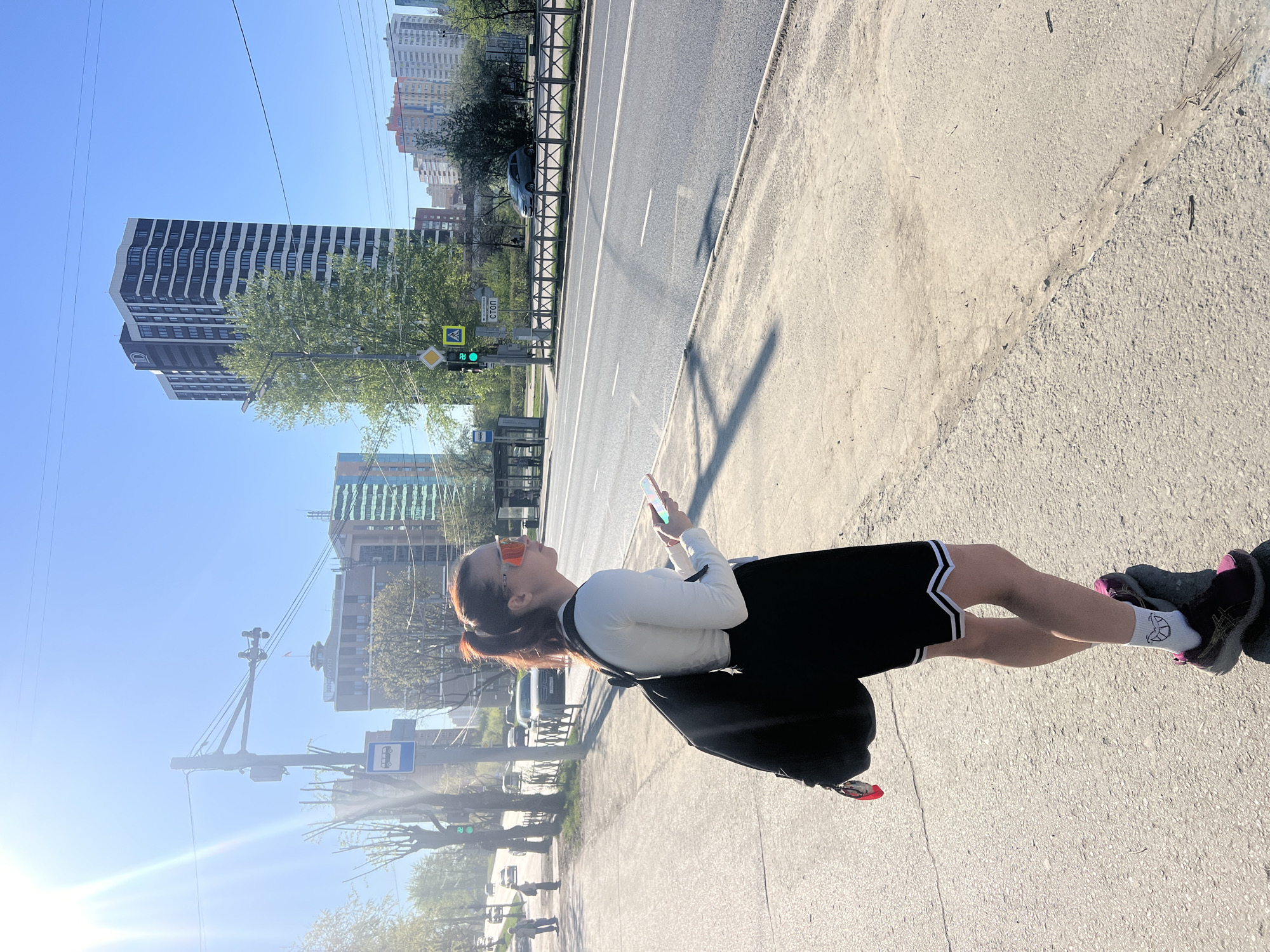 A young woman stands on a sidewalk next to a road in Perm, Russia, holding a smartphone. She wears a white long-sleeved shirt, black athletic shorts with white trim, white socks with a design, and dark sneakers with pink accents. She has a dark backpack over her shoulder and orange sunglasses on. The road is adjacent to the sidewalk with buildings and trees in the background. A tall building is visible in the background to the left. There are streetlights and traffic signals present. The sky is blue and clear.