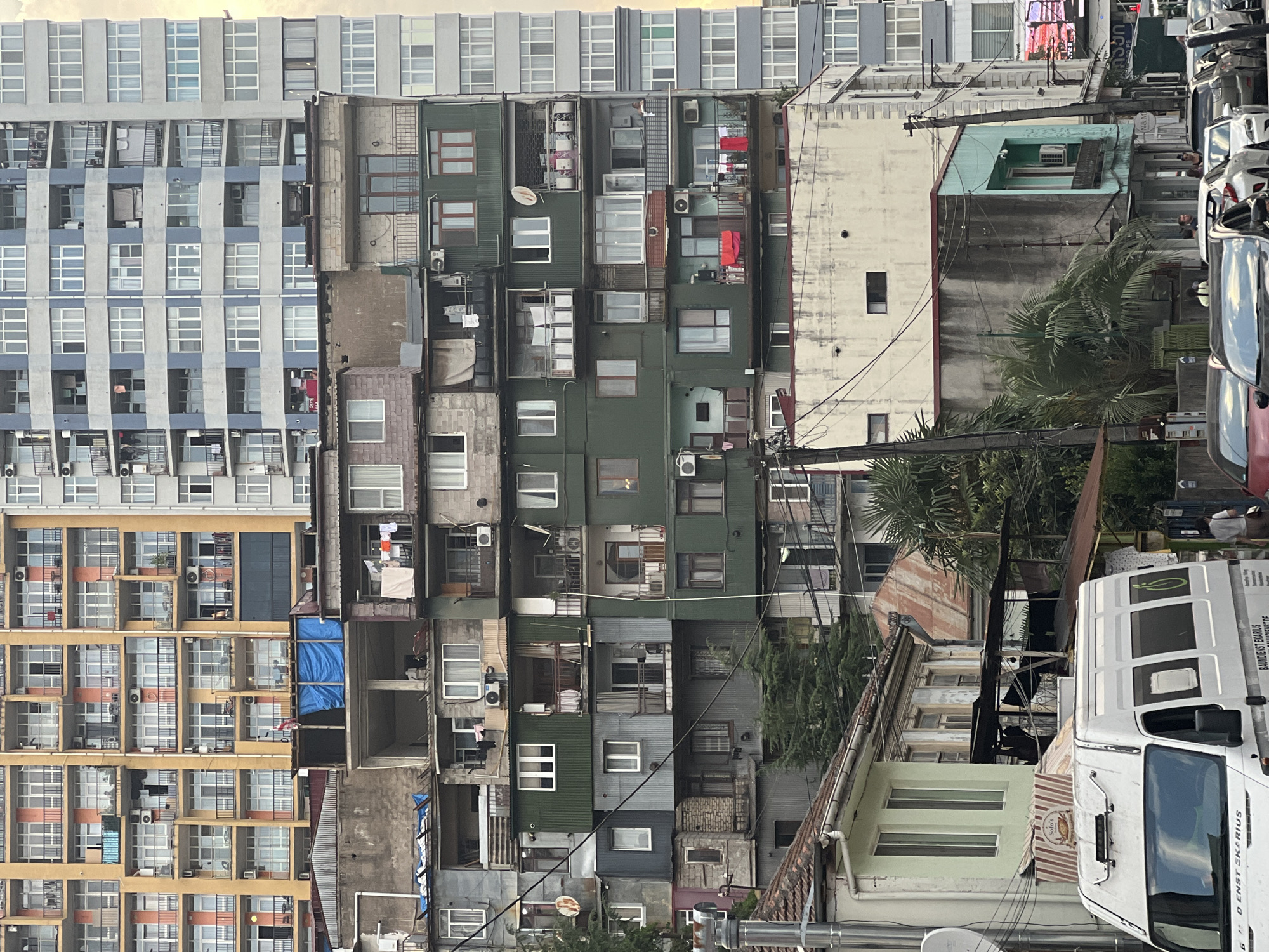 The image depicts a dense urban streetscape in Batumi, Georgia, characterized by a layered composition of buildings with varying architectural styles and states of repair. In the foreground, a white van is partially visible, along with several parked cars. The mid-ground features a collection of older, multi-story buildings with visible signs of wear, including exposed concrete, corrugated metal siding, and makeshift additions. Laundry is hung on balconies, and air conditioning units are mounted on exterior walls, indicating residential use. The background is dominated by several much taller, more modern apartment buildings, one painted in pale yellow and orange hues with repetitive balconies, and another in shades of blue and grey. The overall impression is one of urban density and a mix of old and new construction, with the older structures appearing somewhat dilapidated in contrast to the newer high-rises. The sky is overcast, contributing to a muted, slightly hazy lighting condition.