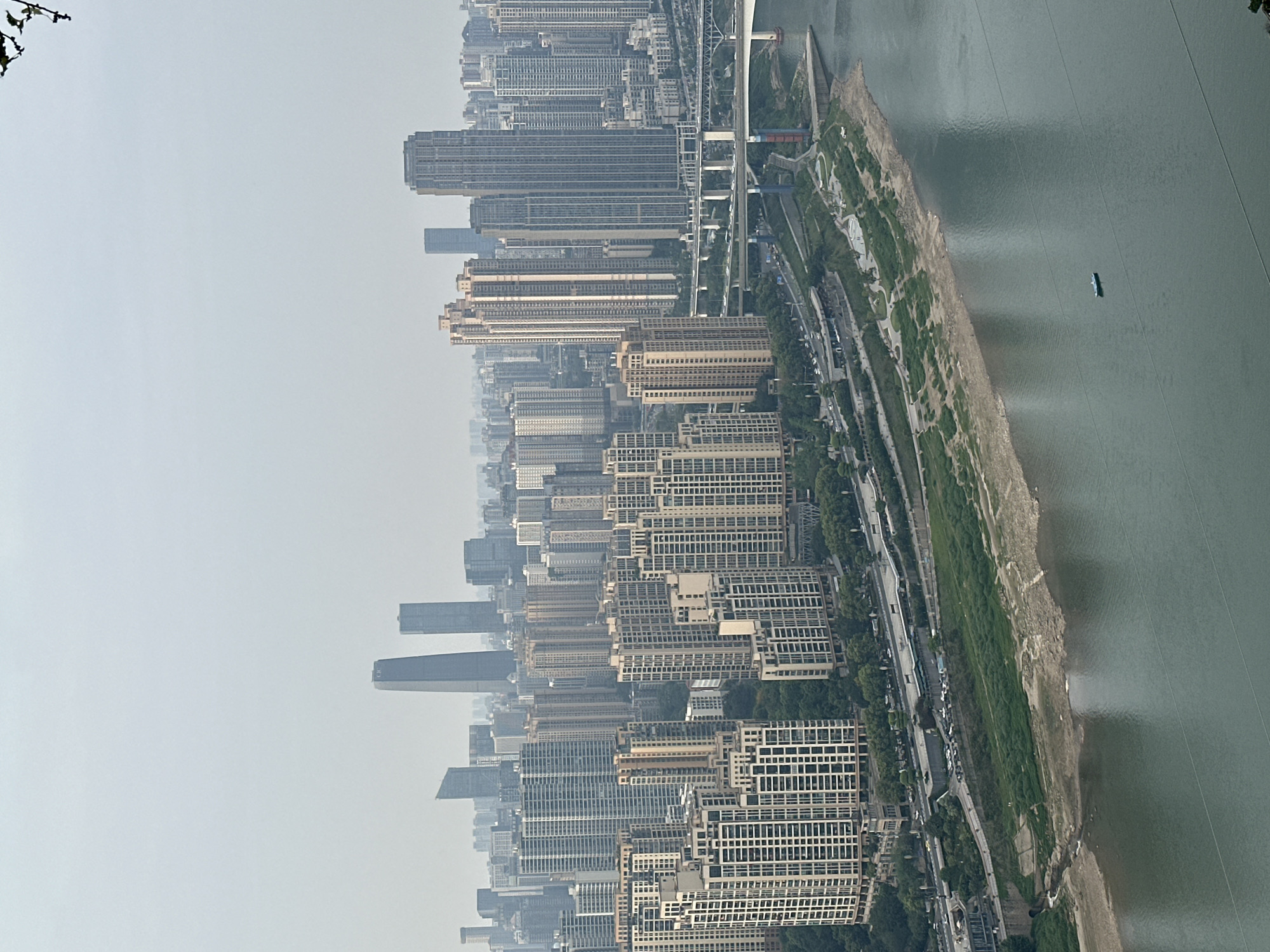 The image shows an aerial view of a densely populated urban area in Yuzhong District, China. The scene is dominated by numerous high-rise buildings, mainly residential apartments, densely packed along the riverbank. A wide river flows through the city. Along the river is an island with a road following the shoreline. A bridge connects the island to the city. The sky is overcast and hazy.