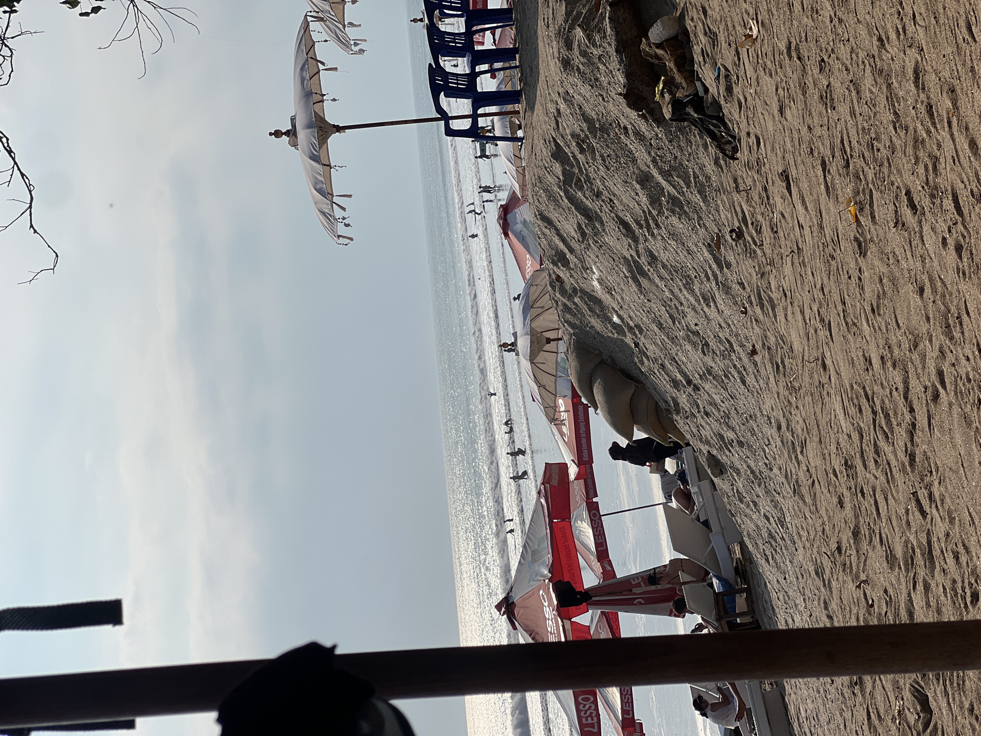 This is a photograph of Kuta Beach in Kuta, Indonesia. The scene is viewed from a sandy embankment with a wooden pole in the foreground, obscuring part of the left side of the image. In the background, the ocean stretches to the horizon, with gentle waves and several people surfing or wading in the water. Along the shoreline, there are numerous beach umbrellas, some red and white with "LESSO" branding, and others white with decorative tassels. Beach chairs are visible under some of the umbrellas, and a few people are seated or lying on them. Further up the embankment, there are sandbags. The sky is overcast with bright sunlight.
