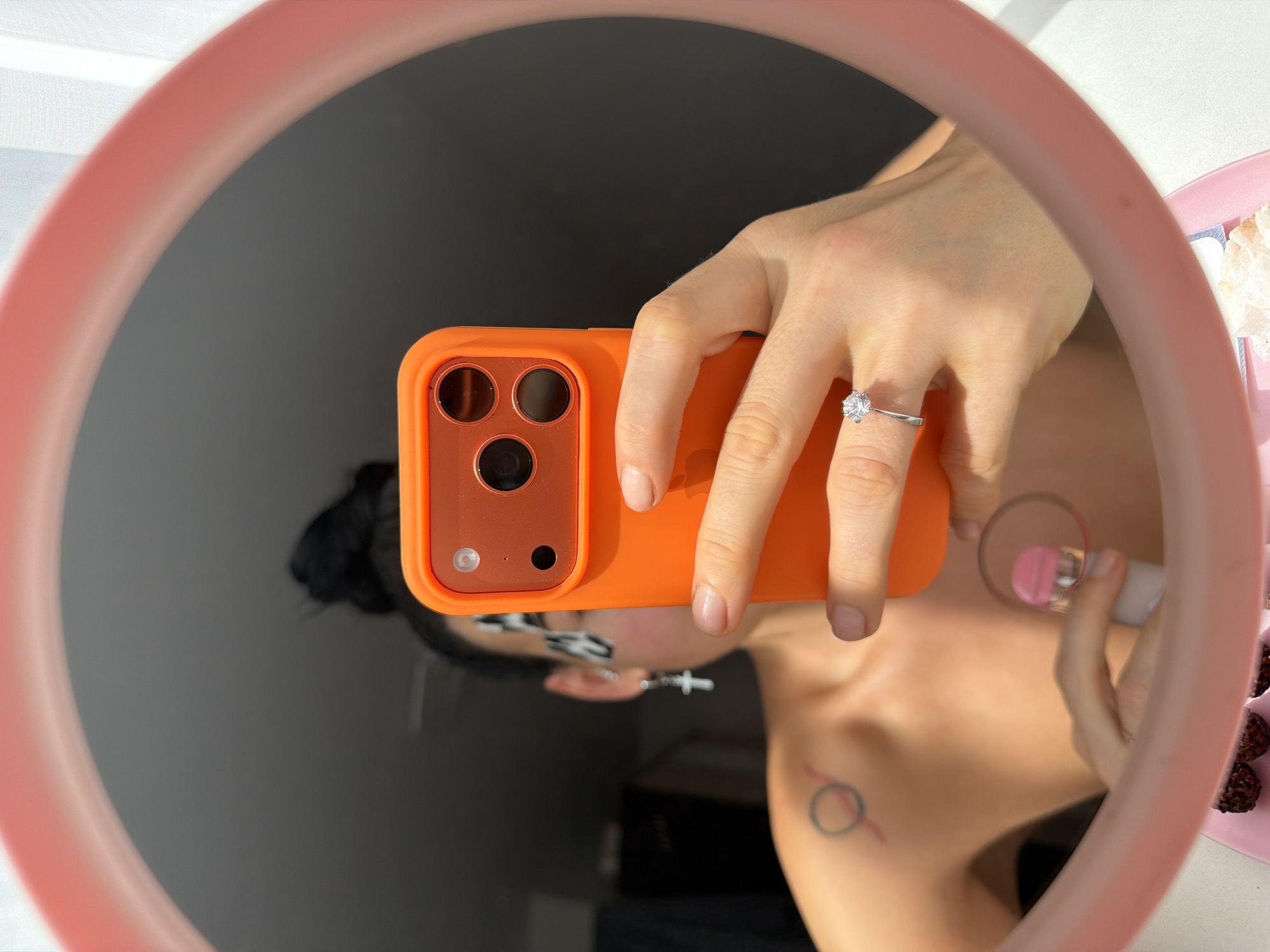An individual is captured taking a mirror selfie, with their reflection appearing inverted in the circular mirror. The person's left hand, adorned with a silver ring featuring a prominent round, clear gemstone, holds an orange-cased smartphone, likely an iPhone, towards the mirror.
In the reflection, parts of the person's head with dark hair are visible, along with an ear wearing a silver cross earring. Their shoulder and collarbone area are exposed, revealing a small circular tattoo outlined in red and black. The person's right hand can also be seen holding a pink and silver beauty product, possibly a foundation stick or highlighter, near their chest.
The mirror has a thick, light pink or peach-colored frame. To the right of the mirror, a light pink plate holds several dark, round chocolates, resembling Ferrero Rocher, and some white decorative elements. The scene is brightly lit, suggesting daylight. No discernible text is visible within the image.