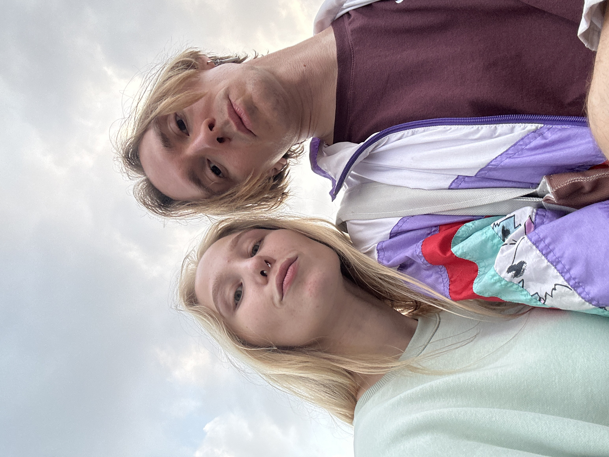An outdoor selfie captures two young individuals from a low-angle perspective, taken against an overcast sky in Budva, Montenegro. The image is rotated such that the subjects appear to be looking down towards the camera.
In the foreground, a young woman with blonde hair and a septum piercing looks directly at the camera with a neutral, calm expression. She is wearing a light mint green top and a vibrant, retro-style windbreaker jacket. The jacket features distinct patches of purple, red, turquoise, and white, with abstract black patterns on some of the white sections.
Behind and slightly above her, a young man with blonde hair and a small silver ear piercing also gazes towards the camera. He wears a dark maroon t-shirt and a multi-colored windbreaker jacket, similar in style to the woman's, with visible purple and white panels.
The background consists of a cloudy, light grey sky, suggesting an overcast daytime. Both individuals' upper torsos and faces are prominently featured in the frame. No text is visible in the image.