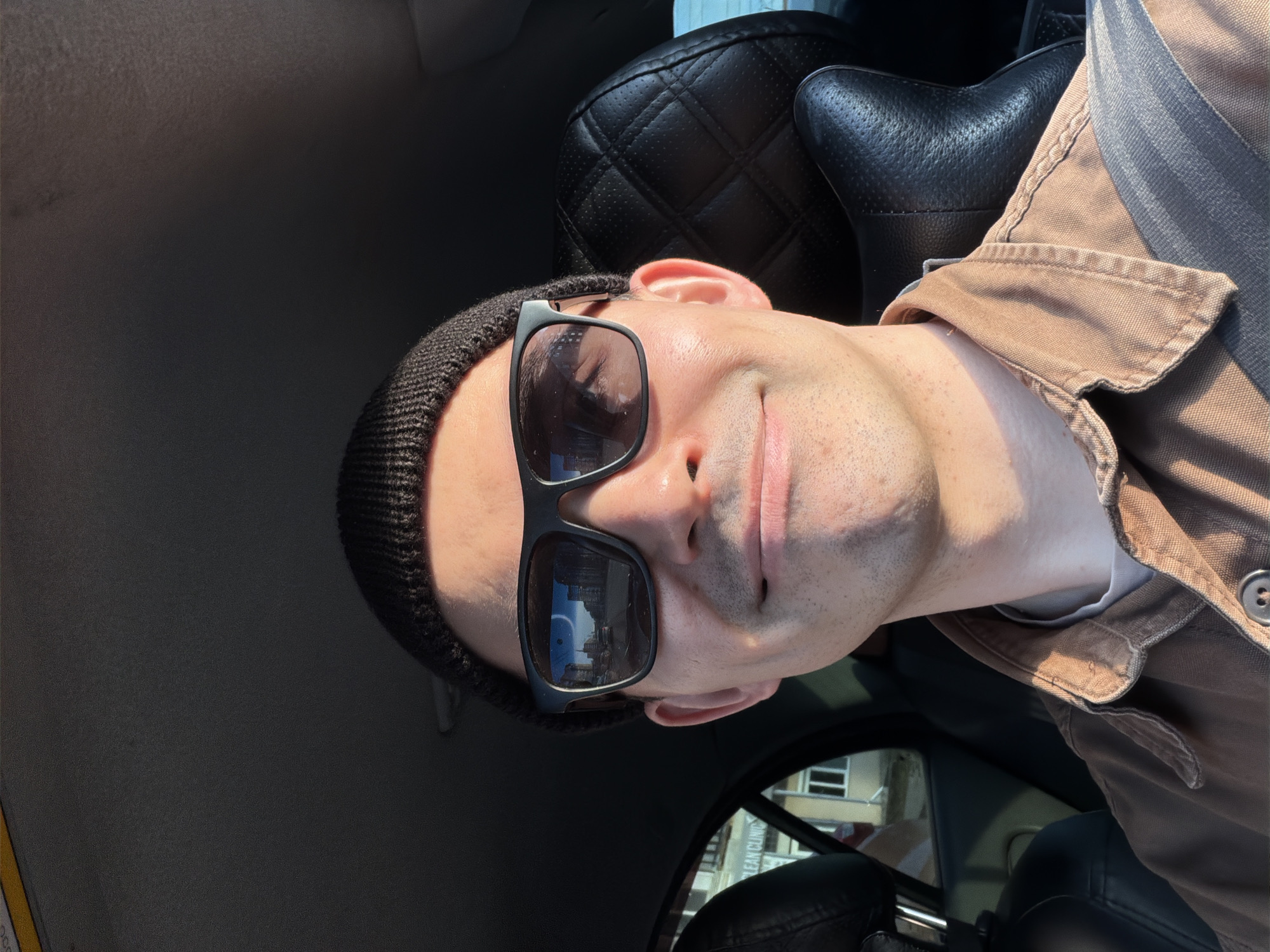 A man wearing sunglasses and a beanie is in a car. He is wearing a brown jacket and a seatbelt. Reflected in his sunglasses are buildings, possibly in Izhevsk, Russia, based on the reversed sign mentioning a "CLEAN CLINIC." The car's interior is visible, including a quilted leather headrest.