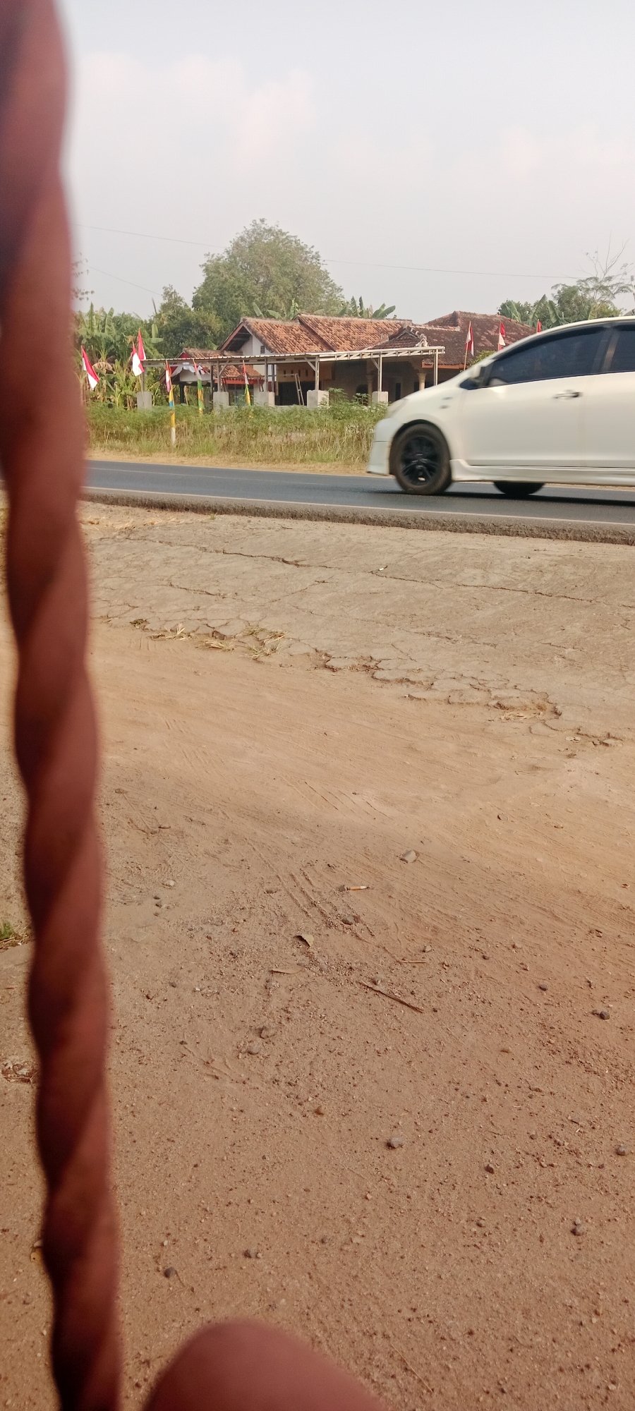The image depicts a roadside scene in Gondang Rejo, Indonesia. In the foreground, a blurred, twisted brown object, possibly a rope or a piece of wood, occupies the left side of the frame. The ground in the immediate foreground is sandy and cracked, with some scattered debris. Beyond this, a paved road runs horizontally across the image. A white car is captured in motion, driving on the road, with its wheels blurred due to the speed. On the other side of the road, there are several houses with red-tiled roofs and traditional Indonesian architectural elements. Small Indonesian flags are visible in front of the houses, suggesting a celebratory occasion or a patriotic display. Lush green vegetation, including banana trees, surrounds the houses. The sky is overcast and hazy, typical of a warm day. The overall scene conveys a rural or semi-rural environment in Indonesia.