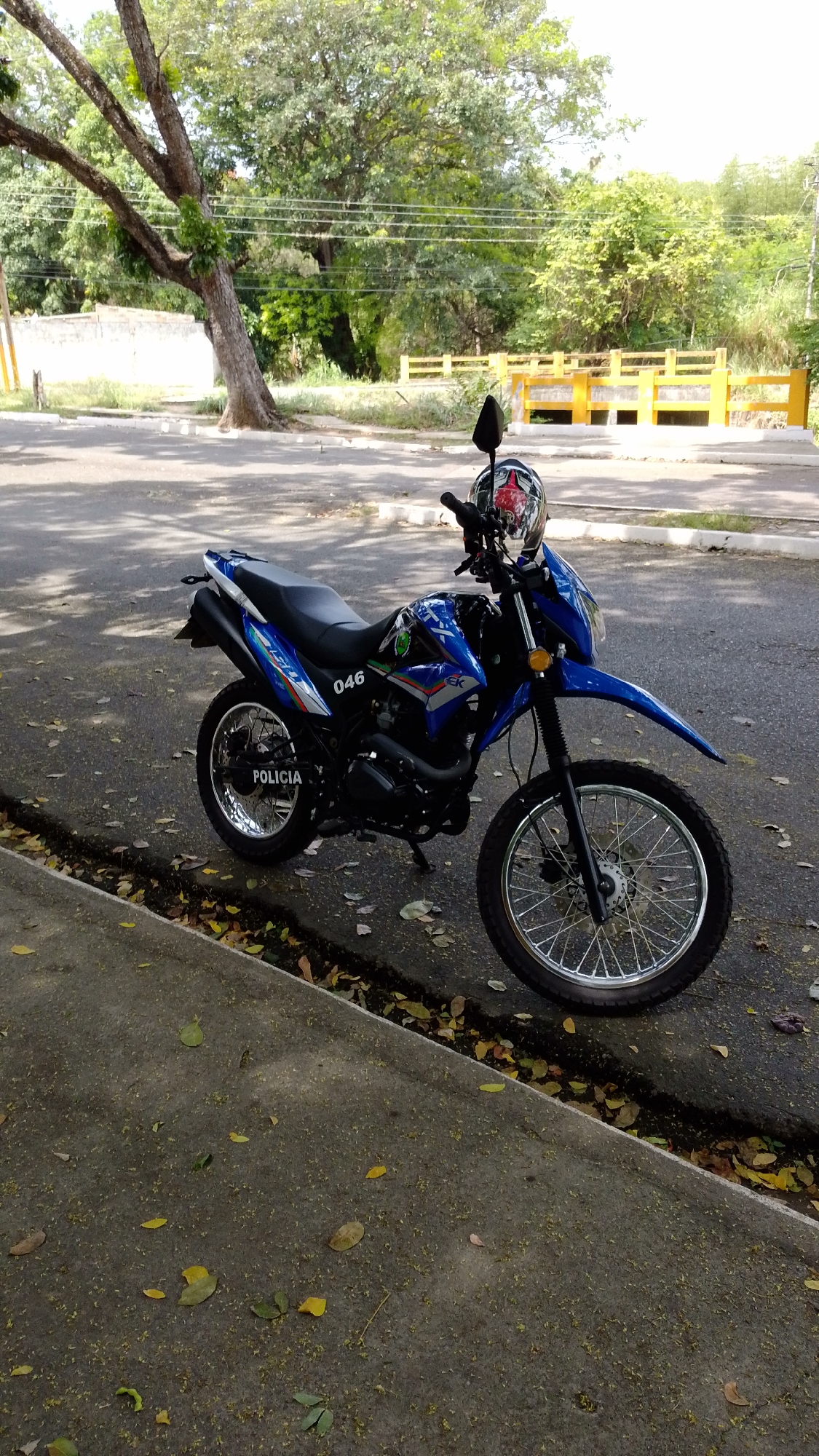 A blue and black police motorcycle is parked on an asphalt road next to a concrete sidewalk in Tinaquillo, Venezuela. The motorcycle, resembling a dirt bike or dual-sport model, features blue front and rear fenders, fuel tank, and side panels, contrasted with a black engine, frame, and seat. The text "POLICIA" is visible in white letters on a black background on the rear side panel, along with the number "046." Additional branding "EK" and possibly "150" are present on the side. A patterned helmet, primarily red and white with a black visor, rests on the motorcycle's handlebars.
The road and sidewalk are scattered with fallen dry leaves of various colors, suggesting a dry season. In the background, a large tree with a thick trunk and leafy green canopy dominates the left side. Beyond the tree, a light-colored wall is visible. To the right, a bright yellow concrete structure, possibly a bridge or barrier, stands amidst lush green foliage and trees. Faint power lines stretch across the upper background. The scene is brightly lit by natural sunlight, indicating daytime, with shadows cast by the motorcycle and surrounding trees.