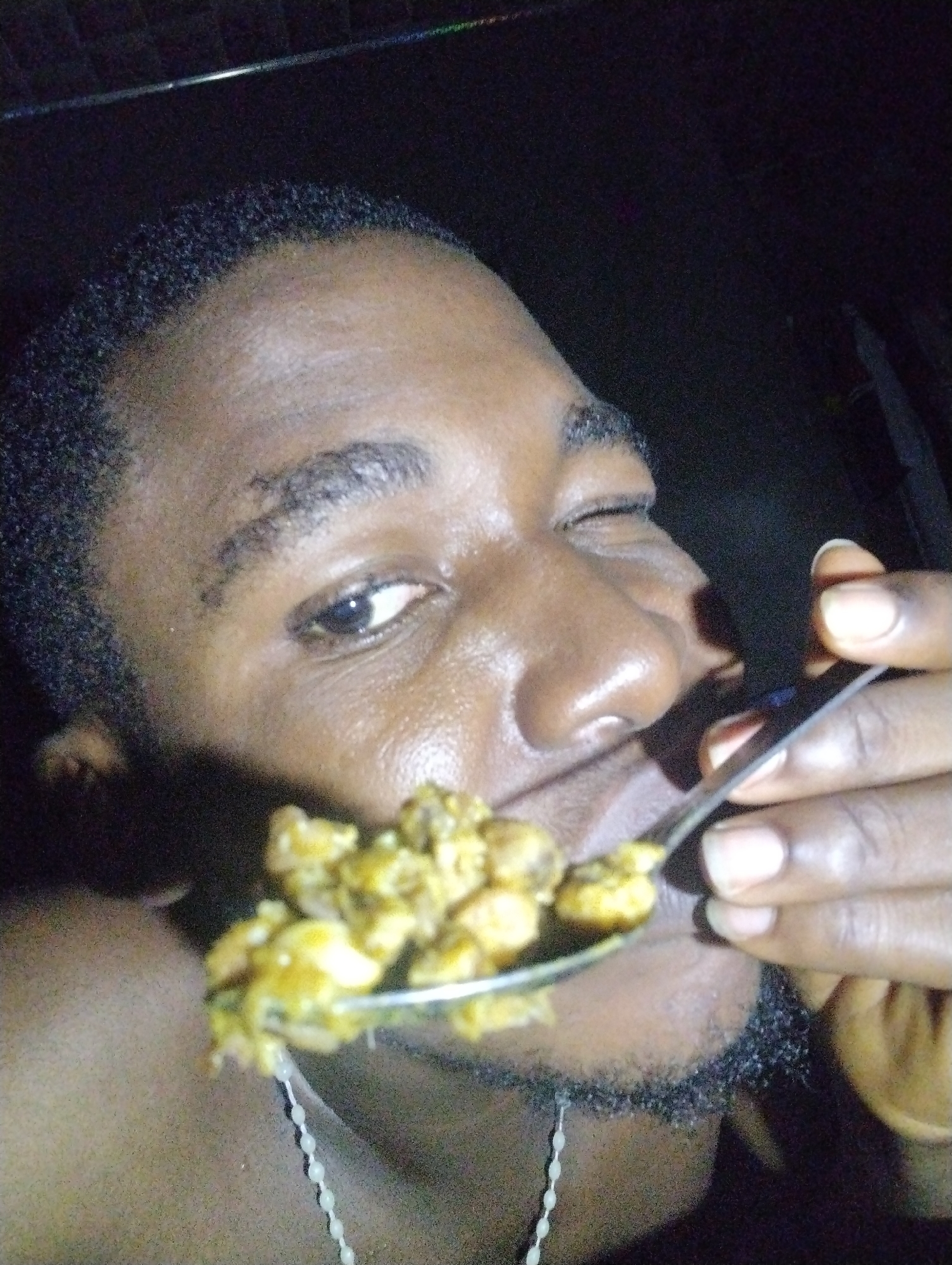 The media file is a close-up, low-light photograph of a Black person with short hair and a beard, eating from a spoon. The person is winking. The food on the spoon appears to be a mixture of small, yellow-brown pieces, possibly chickpeas or similar legumes, in a sauce. Some of the sauce is dripping from the spoon. The setting is dark and appears to be indoors, though no specific details of the location are visible. The lighting is artificial and casts shadows, making it difficult to discern fine details. The overall impression is informal, possibly a candid shot taken during a meal. There is no visible text in the image. The time of day cannot be determined from the image.
