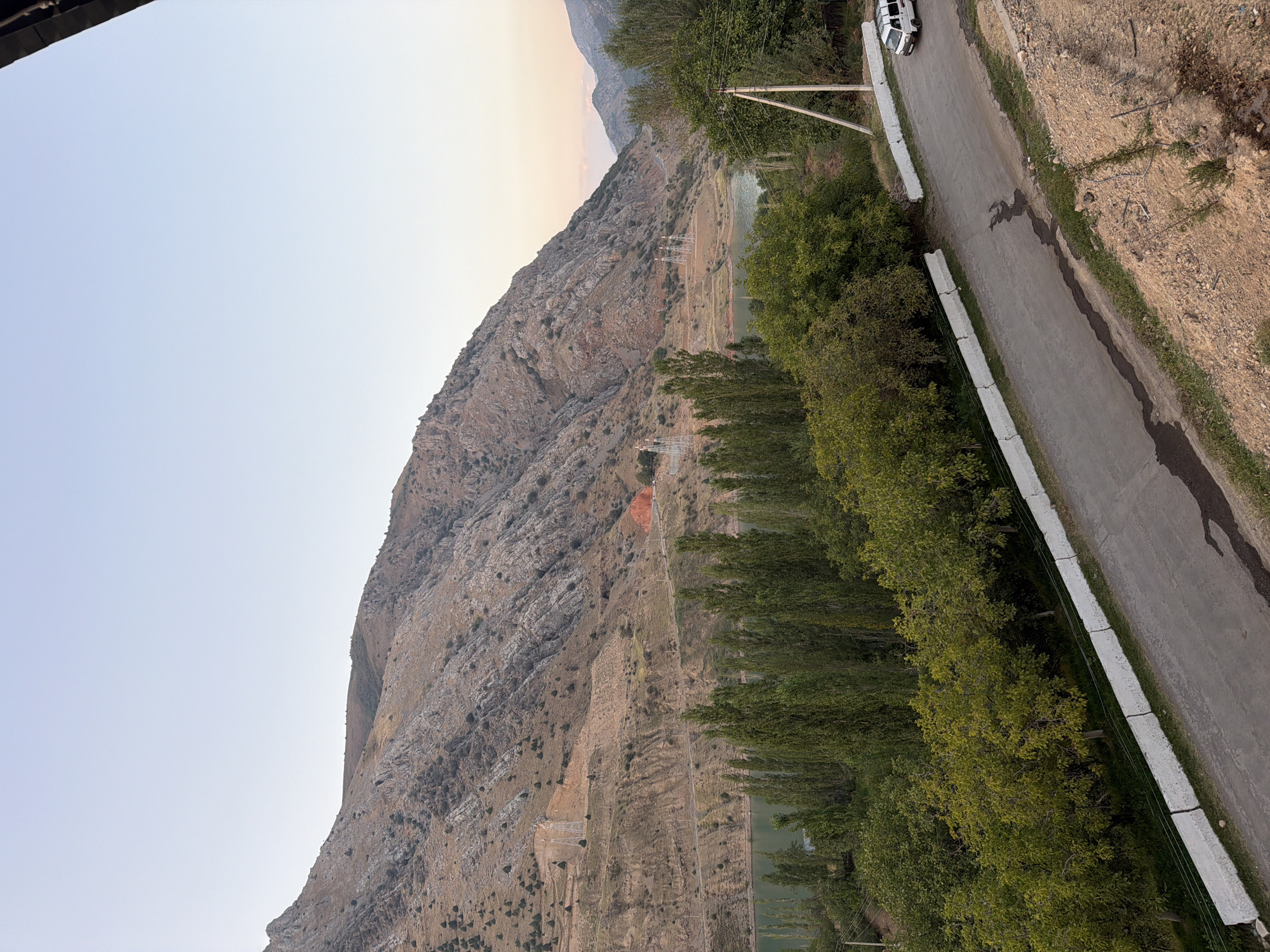 The image depicts a mountainous landscape with a winding road in the foreground, leading to a serene lake bordered by lush green trees. The mountains are rugged and rocky, with sparse vegetation clinging to their slopes. Power lines and pylons are visible in the distance, suggesting human infrastructure within the natural setting. The sky is a pale blue, indicating either early morning or late afternoon. A white van is seen driving on the road, and concrete barriers line the side of the road, separating it from the trees and embankment. The overall scene suggests a rural or semi-rural area, possibly in or near Nanay, Uzbekistan, given the provided location context, characterized by its natural beauty and the presence of a reservoir.