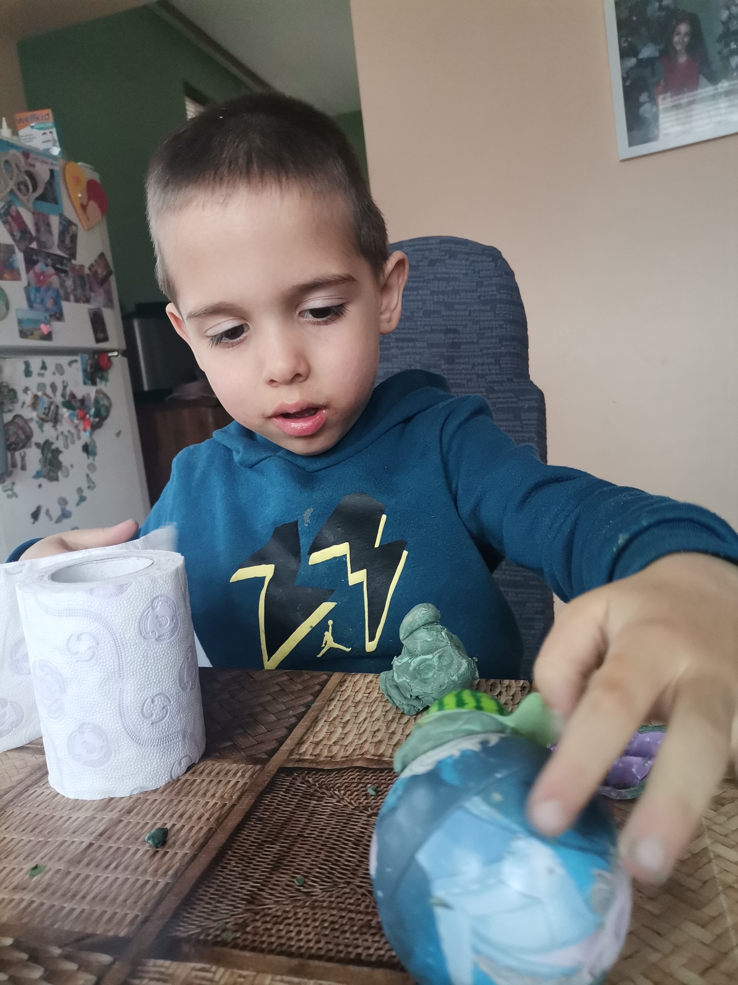 A young boy sits at a table with a woven pattern, crafting with playdough. He wears a blue hooded sweatshirt with a lightning bolt design. In front of him are two rolls of toilet paper, a small green playdough figure, and a blue, white, and purple decorated ball. The background shows a green wall and a refrigerator covered with magnets and pictures. On the right side of the image, a framed photograph hangs on a tan wall. The location is Unknown, Unknown.
