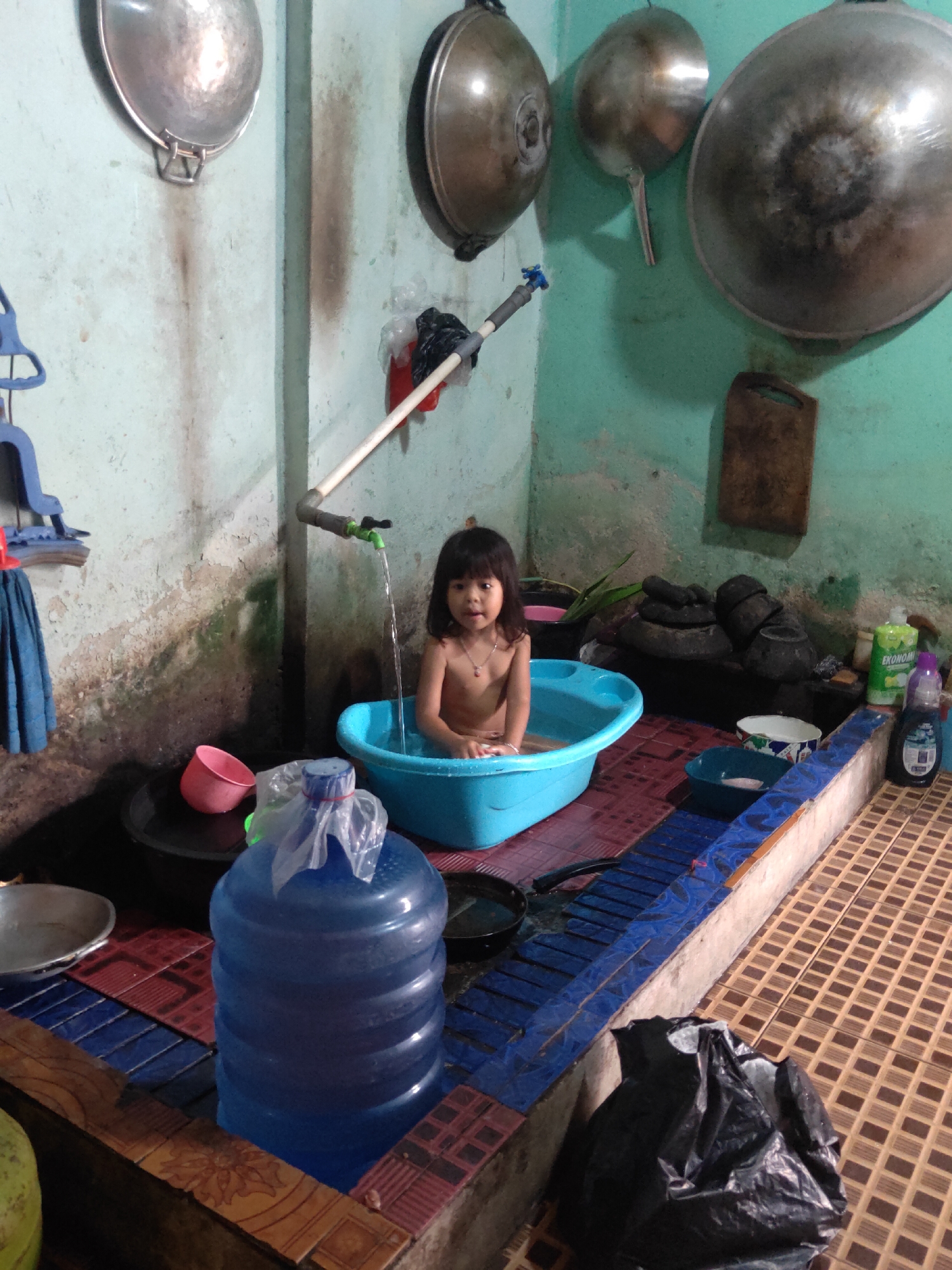 A young child is sitting in a blue plastic bathtub, which is placed on a tiled platform. A faucet overhead is dispensing water into the tub. The child is shirtless and appears to be bathing. The room has a rustic appearance with peeling paint on the walls and what appear to be cooking utensils hanging from the ceiling. In the foreground, a large blue water jug is visible. The scene suggests a modest and functional living space, likely in a home or shared accommodation. The image does not contain any identifiable landmarks or information that would allow for a specific location within Sintang, Indonesia to be determined.