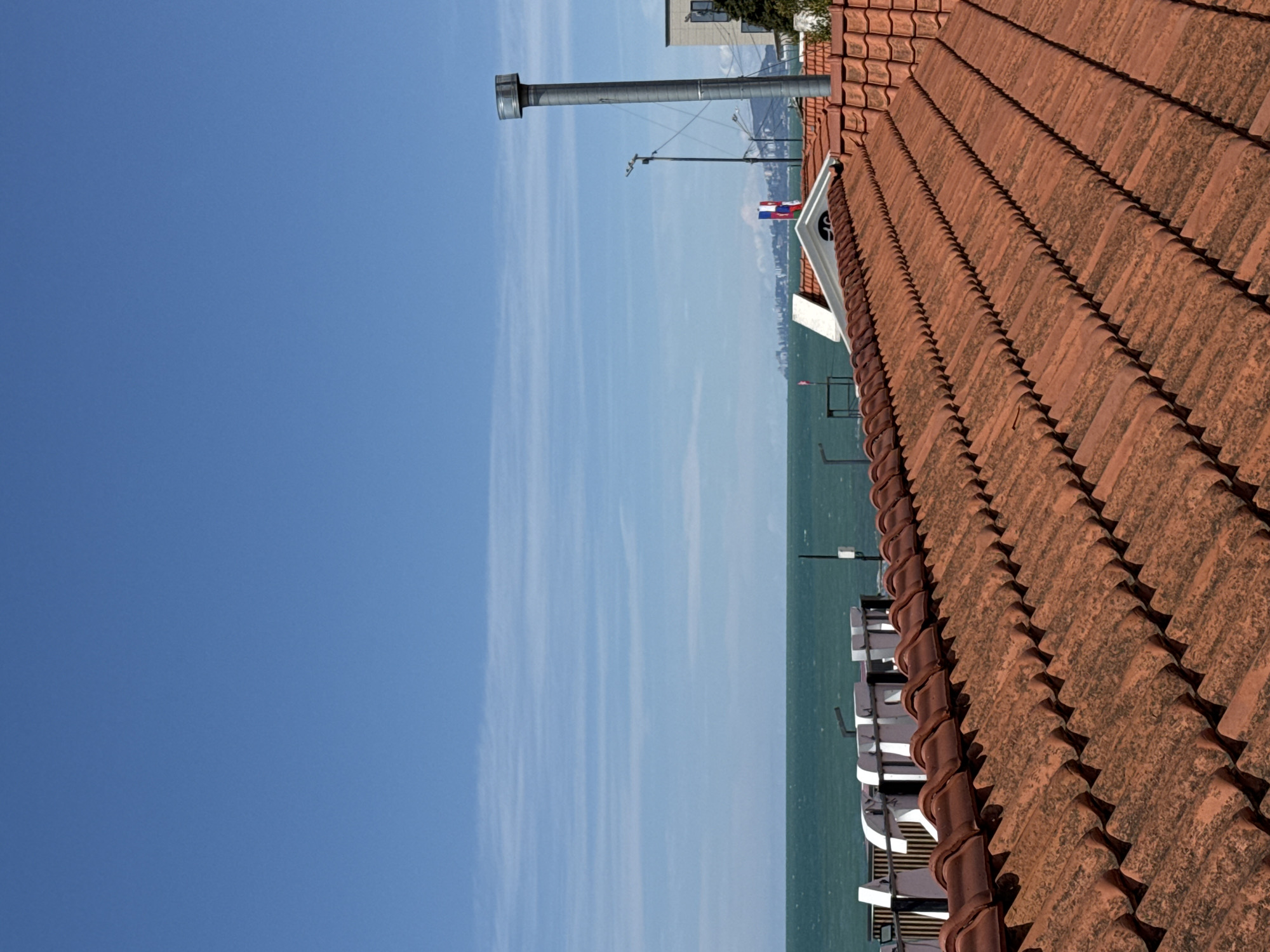 The image shows a view of the sea and sky from a rooftop in Sochi, Russia. In the foreground, there is a section of a red tile roof. A silver chimney and a building are visible behind the roof. The view extends to the sea, with a distant cityscape visible on the horizon. A flag is hanging from a building. The sky is blue, with some white clouds.