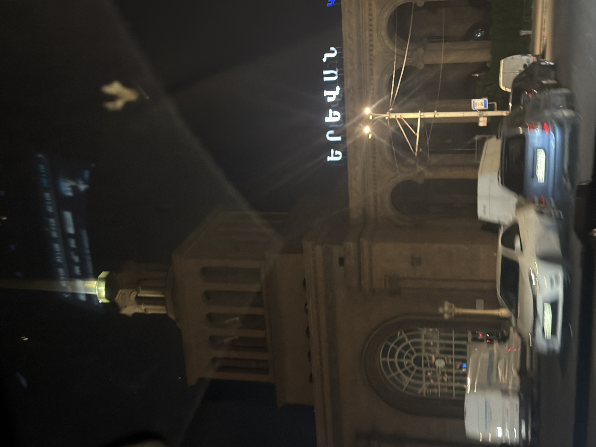 The image captures a nighttime view of the Yerevan Railway Station in Yerevan, Armenia. The station's facade is visible, illuminated by artificial lights. Prominently displayed on the building are the words "ԵՐԵՎԱՆ" in large, white, illuminated letters, spelling out "Yerevan" in Armenian script. A tall, slender spire, lit from below, rises from the building's structure. Several cars are on the street in front of the station, including a white sedan and a blue SUV, appearing to be in motion or stopped in traffic. The street is paved, and there are streetlights illuminating the area. The reflection of lights and possibly the sky is visible on the windows of the station. The overall atmosphere suggests a busy urban environment at night.