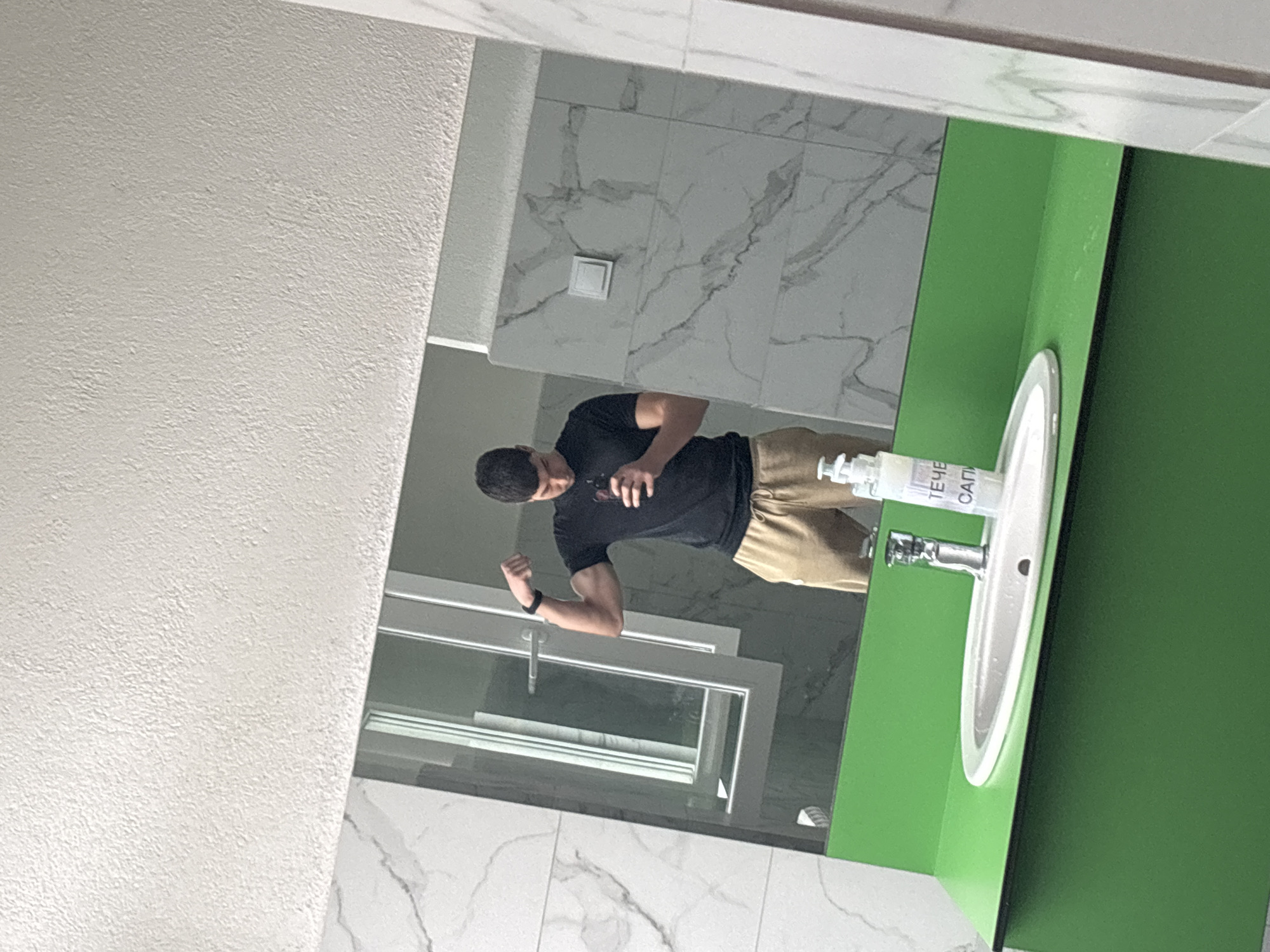 The image is a reflection in a bathroom mirror, likely inside a building, possibly in Zlatograd, Bulgaria. The mirror shows a man wearing a black t-shirt and khaki pants, flexing his arm. He appears to be holding a phone. The bathroom has white, marble-patterned tiles on the wall. A sink with a green cabinet is visible, along with a bottle of hand soap. A window with multiple panes is also reflected in the mirror.