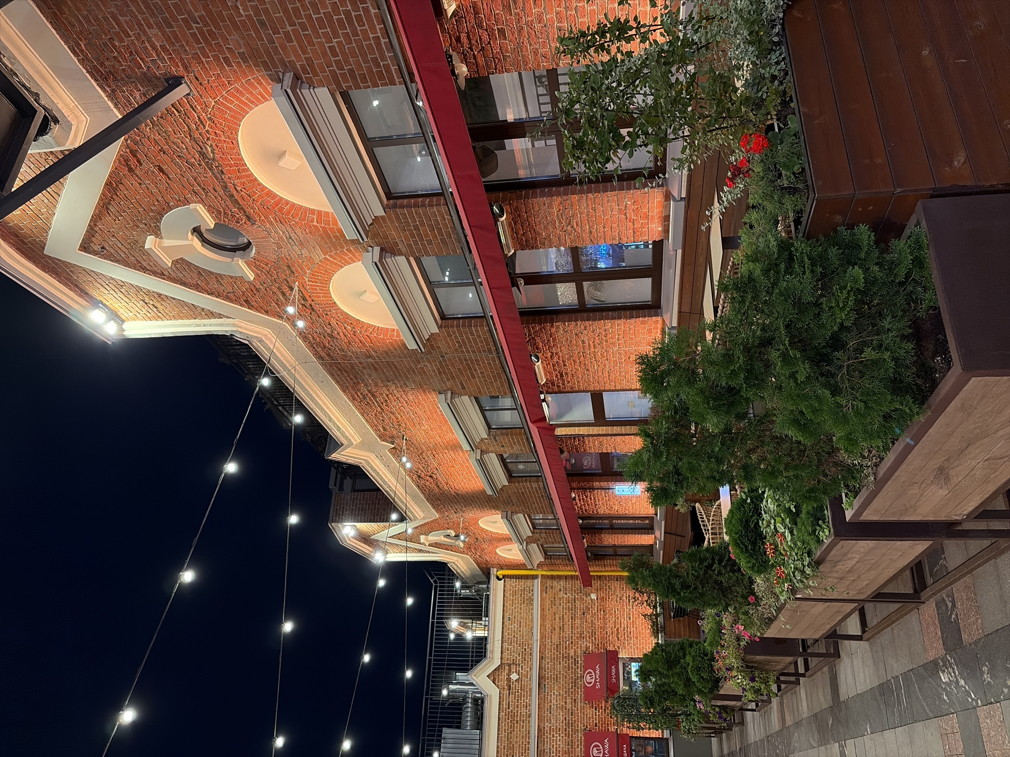 The image captures an outdoor seating area of a restaurant at night in Moscow, Russia. The building is constructed of red brick and features large arched windows, some with red awnings. String lights are strung across the dark sky above. In the foreground, several large wooden planters are filled with greenery and flowers, lining a paved walkway. The overall atmosphere is cozy and inviting, with warm lighting illuminating the building and the plants.