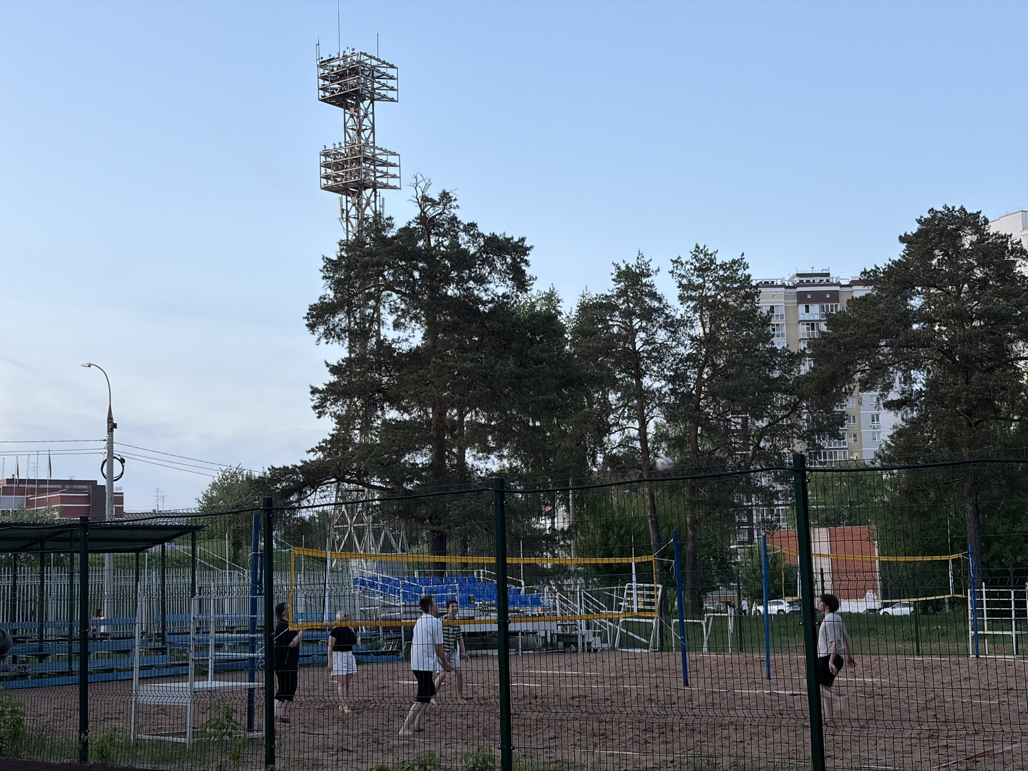 The image depicts a volleyball game in progress on a sandy court in Izhevsk, Russia. The court is enclosed by a metal fence and a net is strung across the center. Six individuals are visible on the court, appearing to be engaged in the game. In the background, there are tall trees, buildings, a metal tower, and streetlights. The sky is light blue, suggesting either early morning or late afternoon lighting.