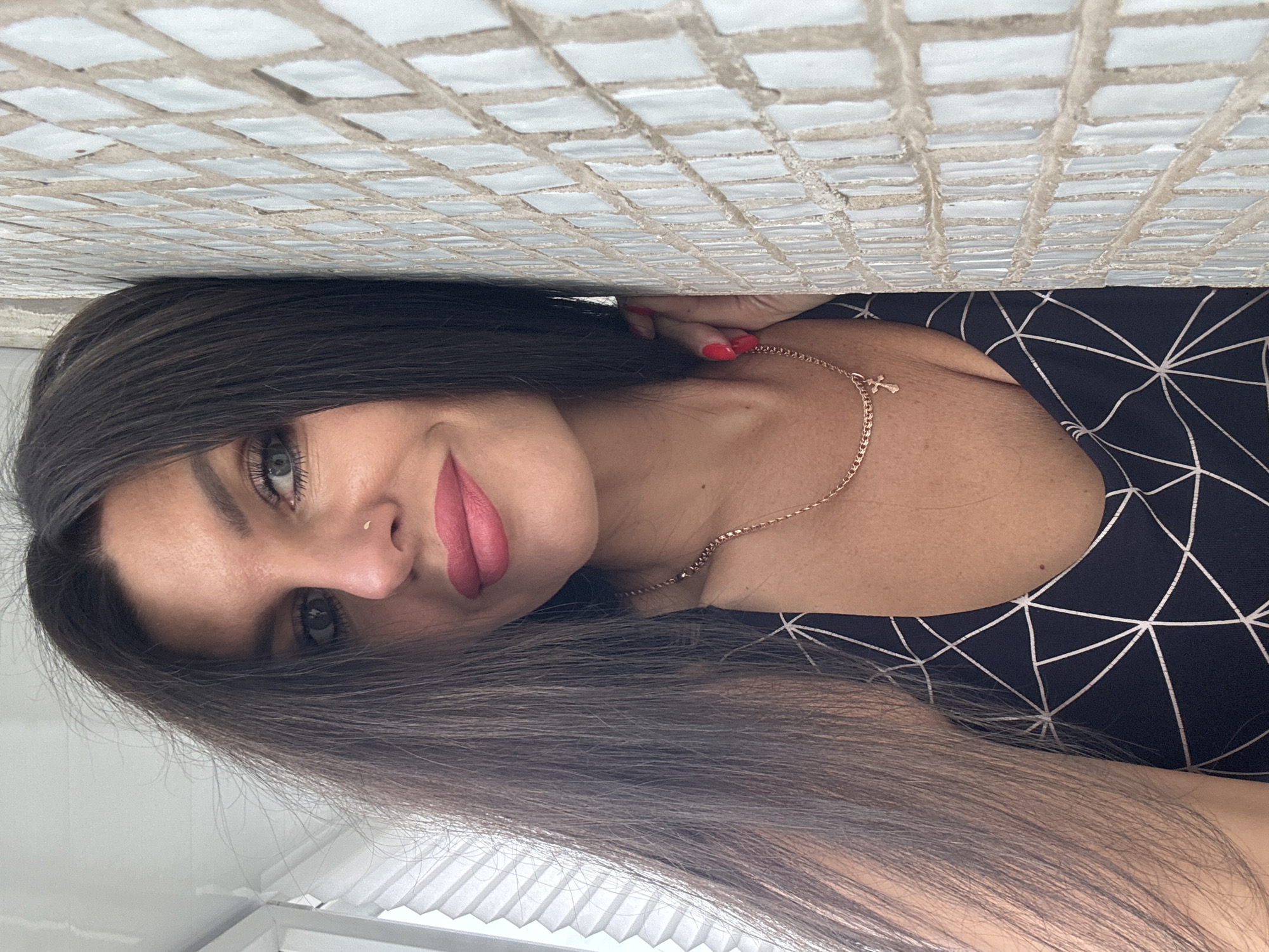 A woman with long, dark hair and bright pink lipstick is leaning against a tiled wall, possibly in an urban setting. She is wearing a dark top with a geometric pattern and a delicate gold necklace with a cross pendant. Her right hand is resting on her neck. The background shows a window with blinds, suggesting an indoor environment. The tiled wall has a mosaic pattern of small, square tiles. No specific location in Chaikovsky, Russia, can be identified from the image.
