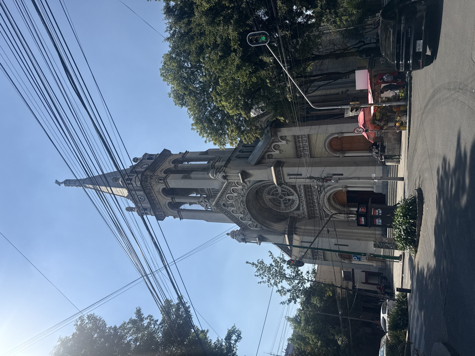 A tilted eye-level shot captures a light-colored, ornate church tower. Power lines crisscross the left foreground, leading up to the tower. Green trees partially obscure the top and sides of the building. A traffic light with a green signal is visible. A black SUV is parked on the right side of the road, with a flower shop nearby under a pink awning. The sky is a clear blue.