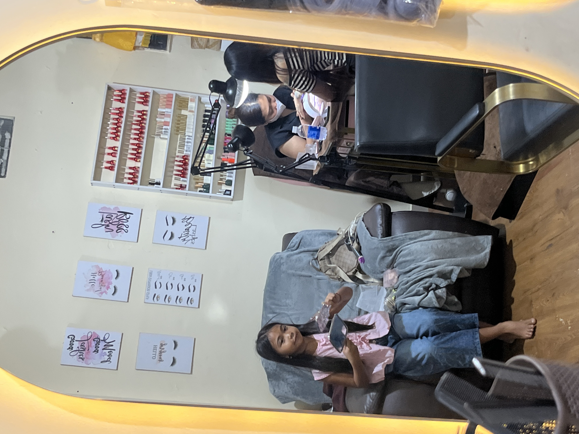 This image depicts the interior of a nail salon, potentially located in North Kuta, Indonesia, as suggested by the context. In the foreground, a young girl with long, dark hair sits on a dark brown leather armchair, draped with a gray blanket. She is wearing a pink and white striped sleeveless top and blue jeans. The girl is holding a small, clear object in her left hand and looking at a small mirror in her right hand. Beside her on the chair is a light brown designer handbag.
In the background, two women are seated at a table, engaged in nail services. One woman, wearing a black top and a white face mask, is attending to the nails of the other woman, who has dark hair and is wearing a black and white striped top. A professional nail lamp is positioned between them. A bottle of water and other small items are on the table.
Behind the table, a white shelving unit displays numerous bottles, likely nail polish, in various colors, predominantly red and pink. The walls are adorned with decorative prints featuring illustrations of eyelashes and calligraphy, suggesting a beauty-themed establishment. The overall lighting appears to be a mix of overhead room lights and task lighting from the salon equipment. The flooring is wooden. A large, oval mirror with a warm, yellow-toned illuminated frame is prominently featured, reflecting parts of the salon. In the bottom left corner, a grey plastic container holds hair combs.