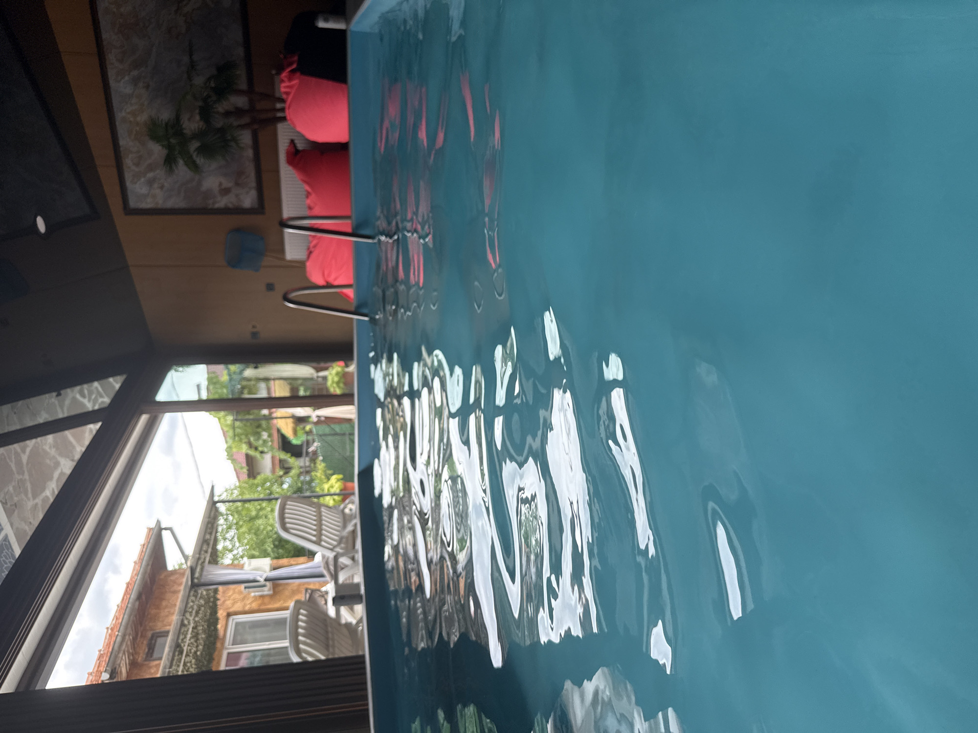 The image depicts an indoor swimming pool with vibrant turquoise water, reflecting the light and creating rippling patterns on the surface. The pool is framed by a glass enclosure, allowing a view of a backyard with a house, lounge chairs, and greenery. In the background, inside the enclosure, are several large, bright pink beanbag chairs and a piece of art on the wall. Two metal pool ladders are visible, partially submerged in the water. The scene suggests a resort or a private residence designed for relaxation and recreation. The location is identified as Chiprovtsi, Bulgaria.