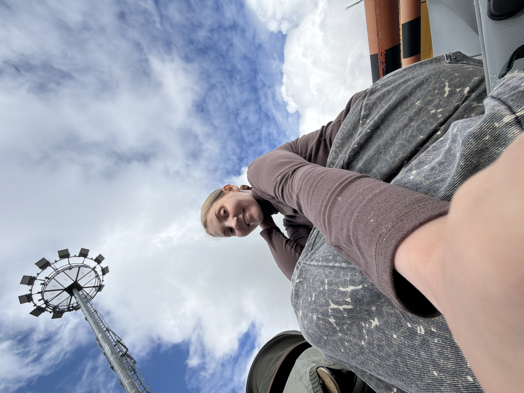 A low-angle shot depicts a young woman with blonde hair, wearing a brown long-sleeved shirt and grey patterned jeans. She is smiling slightly, with her arm extended in front of her as if taking a selfie.
Behind and below her, the scene transitions into a sky filled with blue patches interspersed with white, puffy clouds. A tall metal tower with a circular array of floodlights at the top reaches into the sky. There are some industrial looking equipment where the girl is sitting.
Based on the presence of the distinctive floodlight tower, the location is likely in or around Domodedovo, Russia.