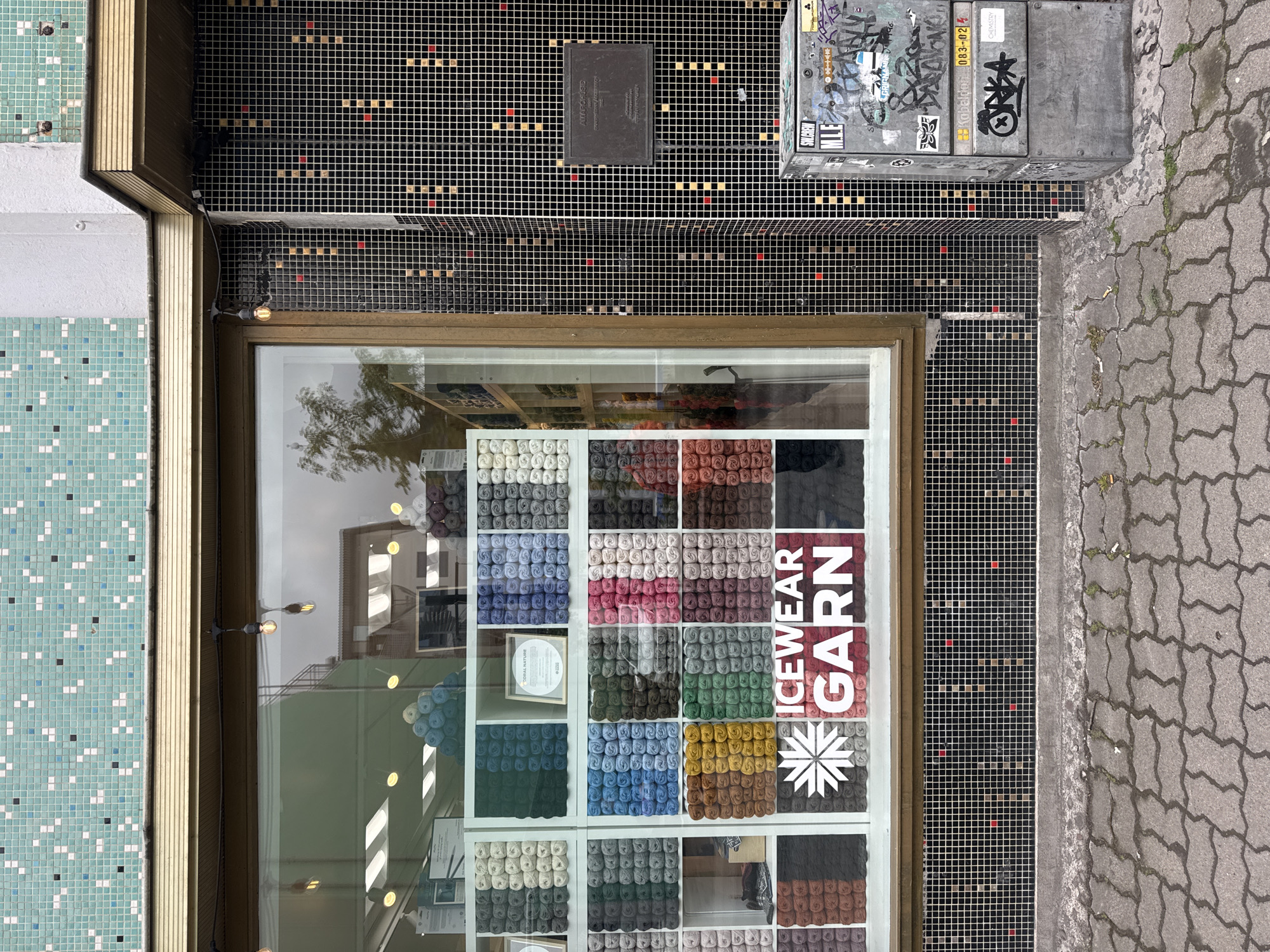 The image captures an overhead perspective of a storefront and its surroundings in Akureyri, Iceland, during daytime. The central focus is a large display window belonging to a shop named "ICEWEAR GARN," indicated by prominent white lettering and a snowflake-like logo on the glass. Inside, numerous skeins of yarn are neatly arranged in cubbies, showcasing a wide spectrum of colors including various shades of white, grey, blue, green, yellow, and red.
Above and to the right of the shop window, the building facade is covered in small, black mosaic tiles, accented with scattered red, beige, and white squares. A rectangular black box, possibly an electrical panel, is mounted on this tiled surface, with faint text visible such as "ATK-ESP". A string of two bare light bulbs hangs above the storefront. To the far left, another section of the building features light blue and green mosaic tiles with white and dark accents.
To the right of the tiled facade, a large, grey, utilitarian box-like structure is visible, covered extensively in graffiti and stickers. Identifiable text on this box includes "SVERKEN", "08.3-02", and "CHEMISTRY". The ground surface adjacent to the building is paved with grey interlocking pavers, exhibiting some wear, cracks, and small weeds. The scene appears still and devoid of people, suggesting a quiet street or alleyway. The lighting indicates daylight, with reflections in the window suggesting a bright, possibly overcast, sky.