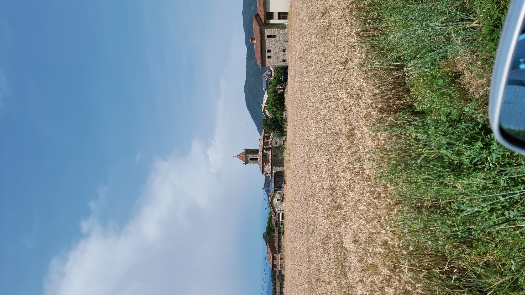 The image shows a rural landscape with a wheat field in the foreground and a village nestled in the background. The wheat field is golden and ripe, suggesting it is late summer or early autumn. In the village, a prominent church with a bell tower stands out. Surrounding the church are several stone houses with red-tiled roofs, characteristic of Spanish architecture. Rolling green hills form the backdrop to the village, under a bright blue sky with scattered white clouds. A car's side mirror is partially visible in the bottom right corner, indicating the photo was taken from inside a vehicle. The scene depicts a tranquil, idyllic view of the Spanish countryside.