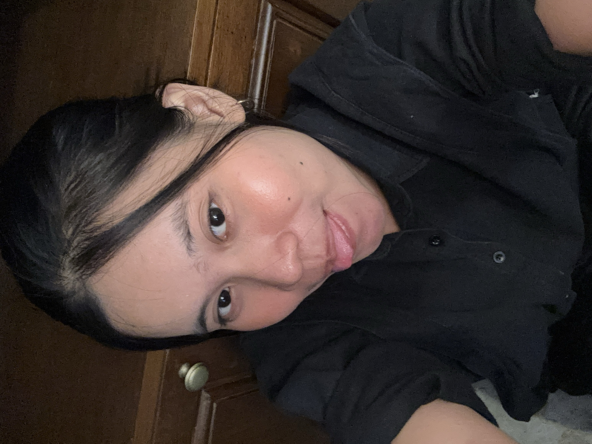 A woman of Asian descent is captured in a close-up, selfie-style photograph. She is wearing a black collared shirt with buttons. Her dark hair is pulled back. Her facial expression is playful, with her tongue sticking out slightly. The background consists of wooden furniture, possibly a cabinet or dresser, with a brass knob visible on the left side. The setting appears to be indoors. No specific location in South Jakarta, Indonesia, can be determined from the image.
