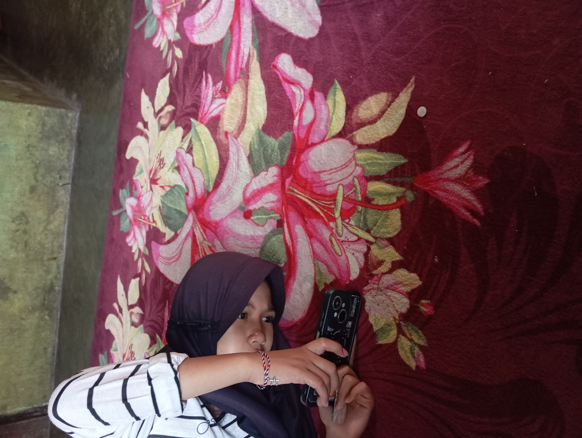 A young female wearing a dark hijab and a black and white striped shirt is lying on a maroon blanket with a floral pattern. She is holding a black smartphone with a case, which is on the blanket. Her left arm is bent, and her hand is resting on the phone. A bracelet with a star charm is visible on her wrist. The blanket is spread on a tiled floor. The background shows a wall with a green textured surface. The scene appears to be indoors, likely in a residential setting in Rejosari, Indonesia.