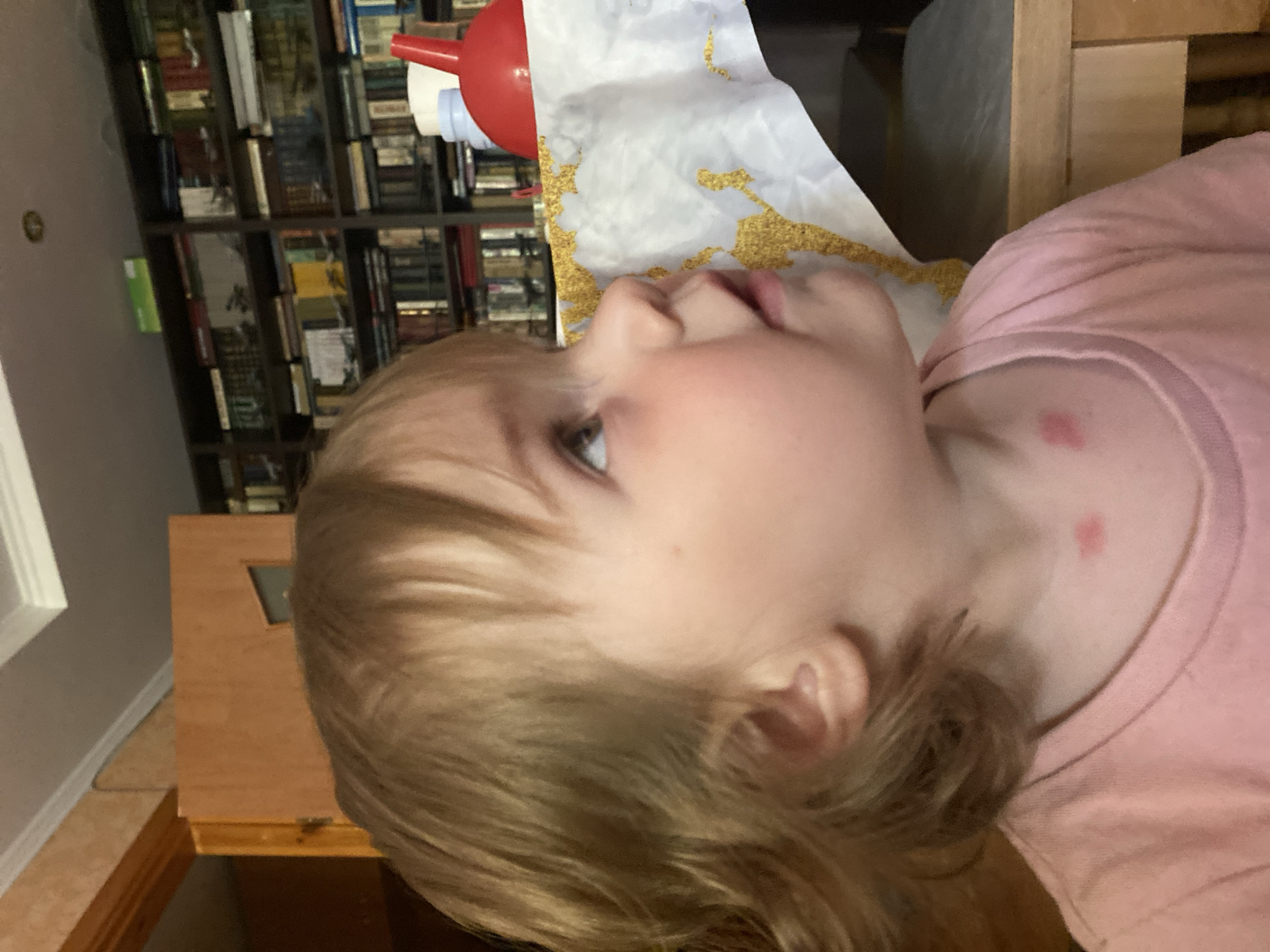 The image shows a close-up, side profile of a young child with blonde hair, wearing a pink shirt. The child's face is turned upwards and to the right, with their mouth slightly open and their eyes looking up. There are two small red marks on the child's neck, one slightly above the other.
In the background, a bookshelf filled with books is visible. To the right of the child's head, a wooden structure, possibly furniture, is present. A white sheet with a gold pattern is draped in the background, near a red and white object that appears to be a toy.
The lighting suggests it could be indoors, possibly in a home environment. The image is a still photograph. No specific time of day or weather conditions are indicated. The overall mood is difficult to ascertain due to the close-up nature of the shot and the child's pose. There is no visible text in the image.