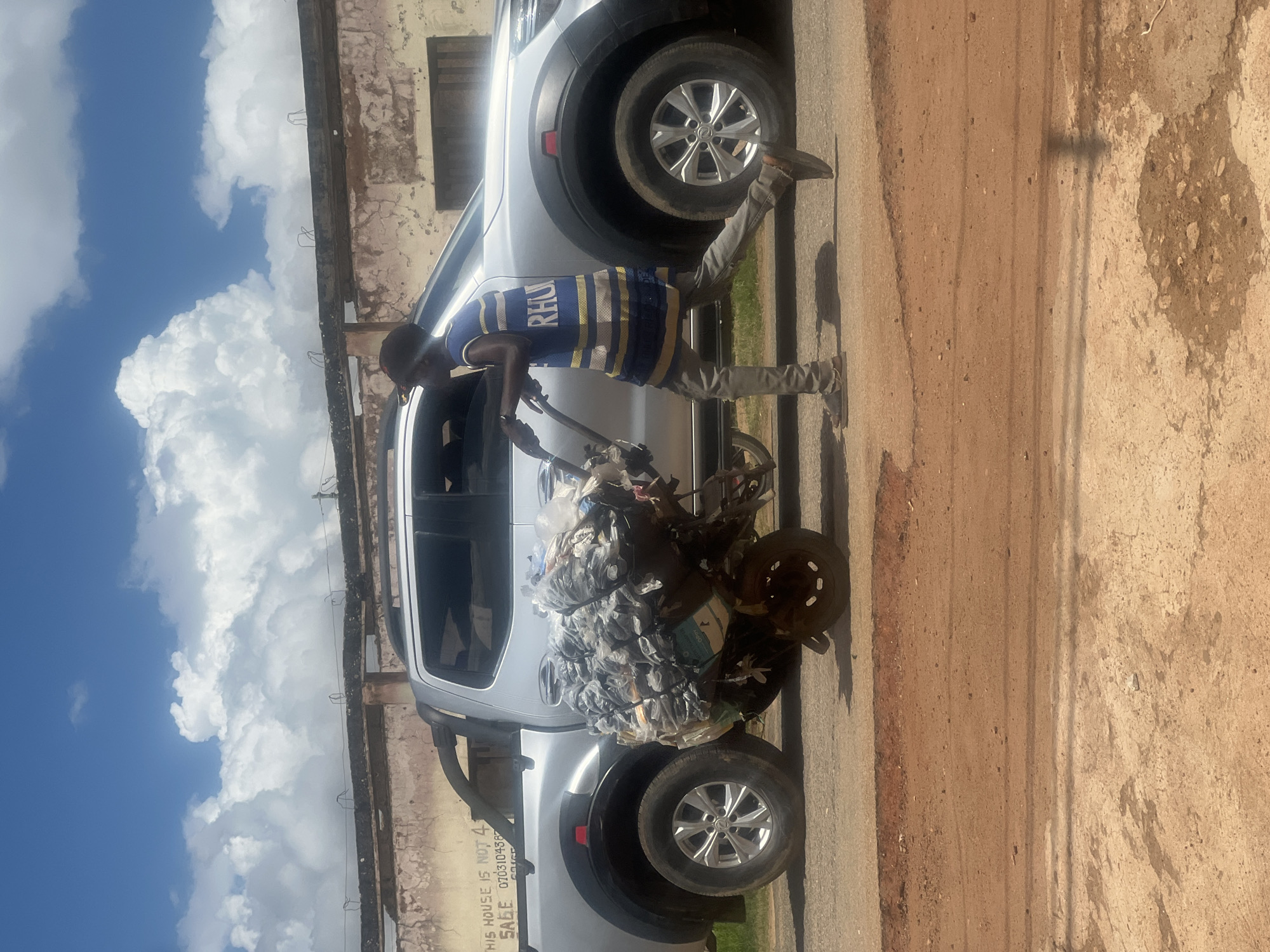 A young boy is pushing a cart overloaded with plastic waste down a dirt road. The boy is wearing a blue and yellow striped shirt and grey pants. He is walking next to a silver pickup truck parked on the side of the road. In the background, a building with peeling paint and a barbed wire fence is visible. The sky is blue with fluffy white clouds. The scene is set in the city of Jos, Nigeria.