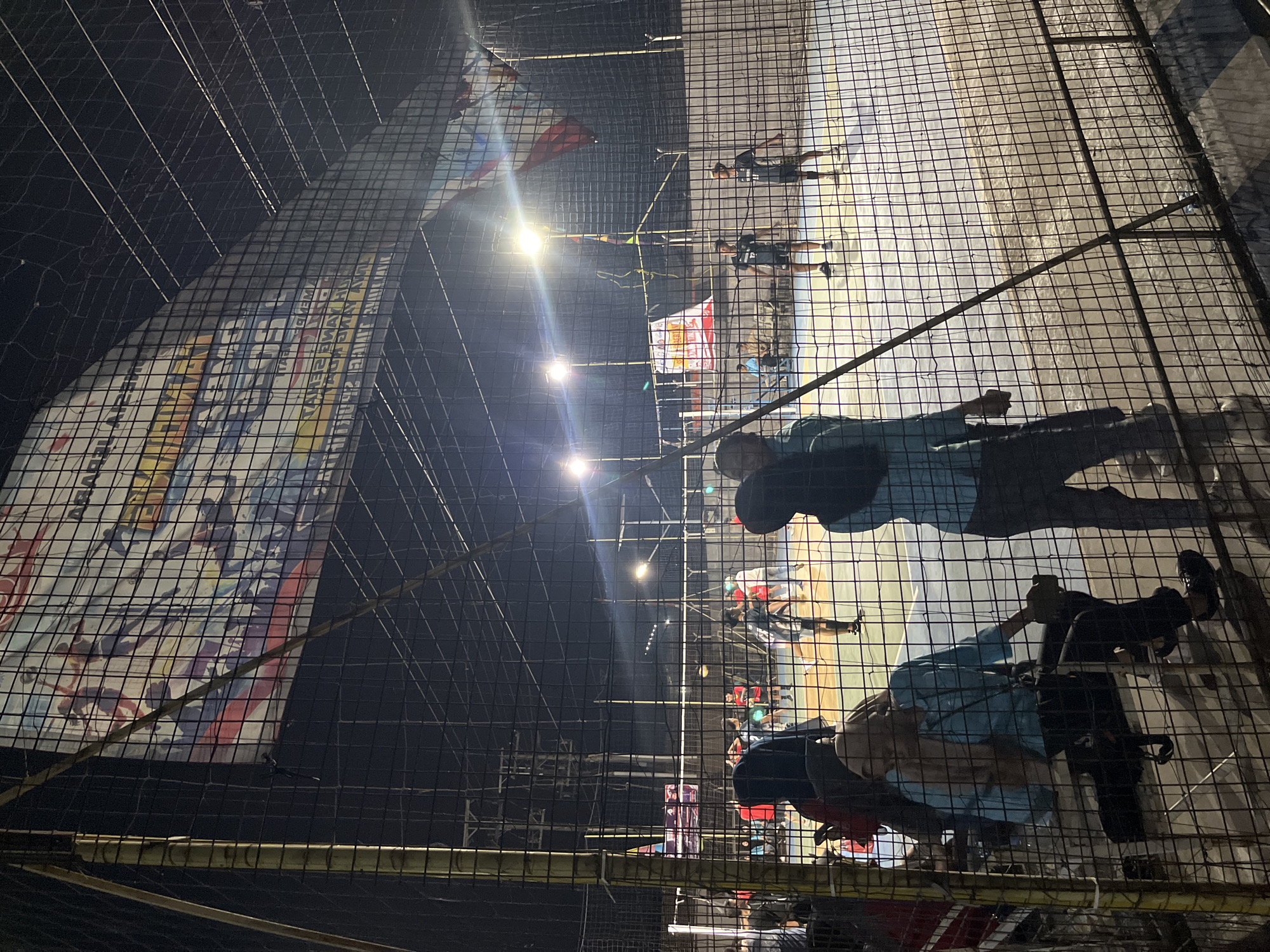 An outdoor volleyball court at night is illuminated by bright floodlights. Several individuals are present, some actively playing volleyball on the court while others are seated or standing near the perimeter fence. The court appears to be made of concrete, and a net divides the playing area. In the background, there are structures including poles and possibly buildings. A large banner with graphics and text is suspended above one end of the court. The scene is viewed through a mesh fence, suggesting the spectators are outside the playing area. The location is identified as Pelang, Indonesia.