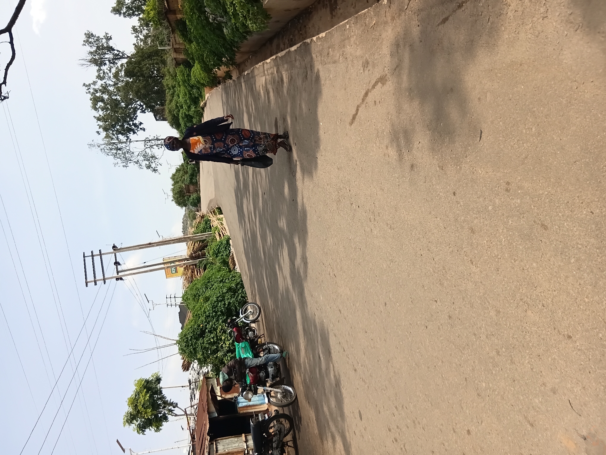 A woman in a long, patterned dress and a dark jacket walks away from the camera on a paved road. She is carrying a dark bag in her left hand. The road is lined with lush green bushes on the right and a few motorcycles parked on the left. Behind the motorcycles is a small, makeshift structure made of corrugated metal. In the background, there are trees, utility poles with electrical wires, and a pile of lumber. The sky is clear and blue. The scene appears to be an urban or semi-urban environment.