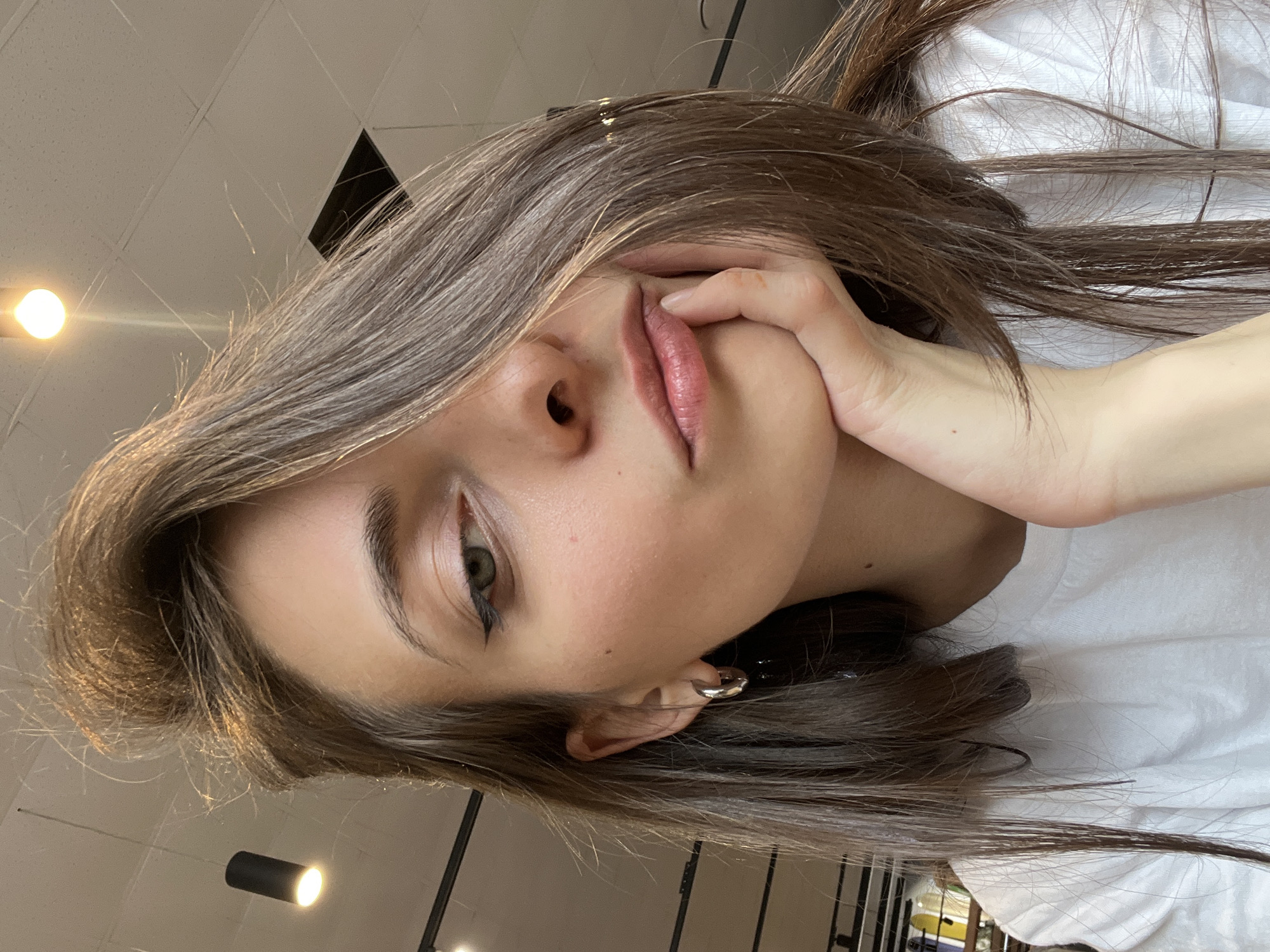 A close-up, low-angle shot shows a woman with light brown hair, wearing a white shirt, and holding her face with her left hand. She has light skin, dark eyebrows, and green eyes. She is wearing silver hoop earrings. The background shows a commercial ceiling with recessed lighting. The location is not specified. There is no explicit indication of Krasnoyarsk, Russia in the image.
