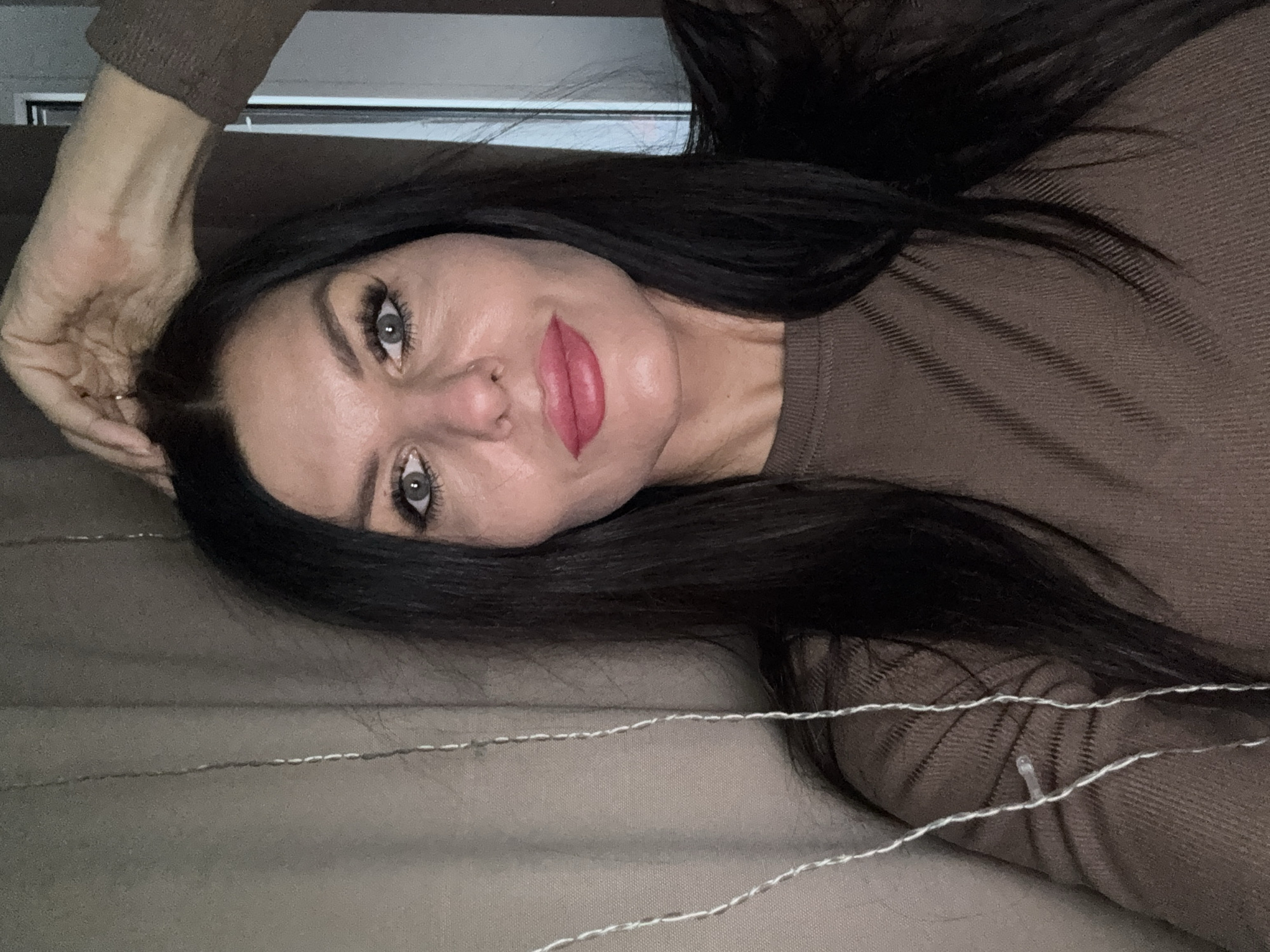A woman with dark hair and fair skin is lying down. She wears a brown long-sleeved shirt. Her hand is near her head. The location is indeterminable from the image.