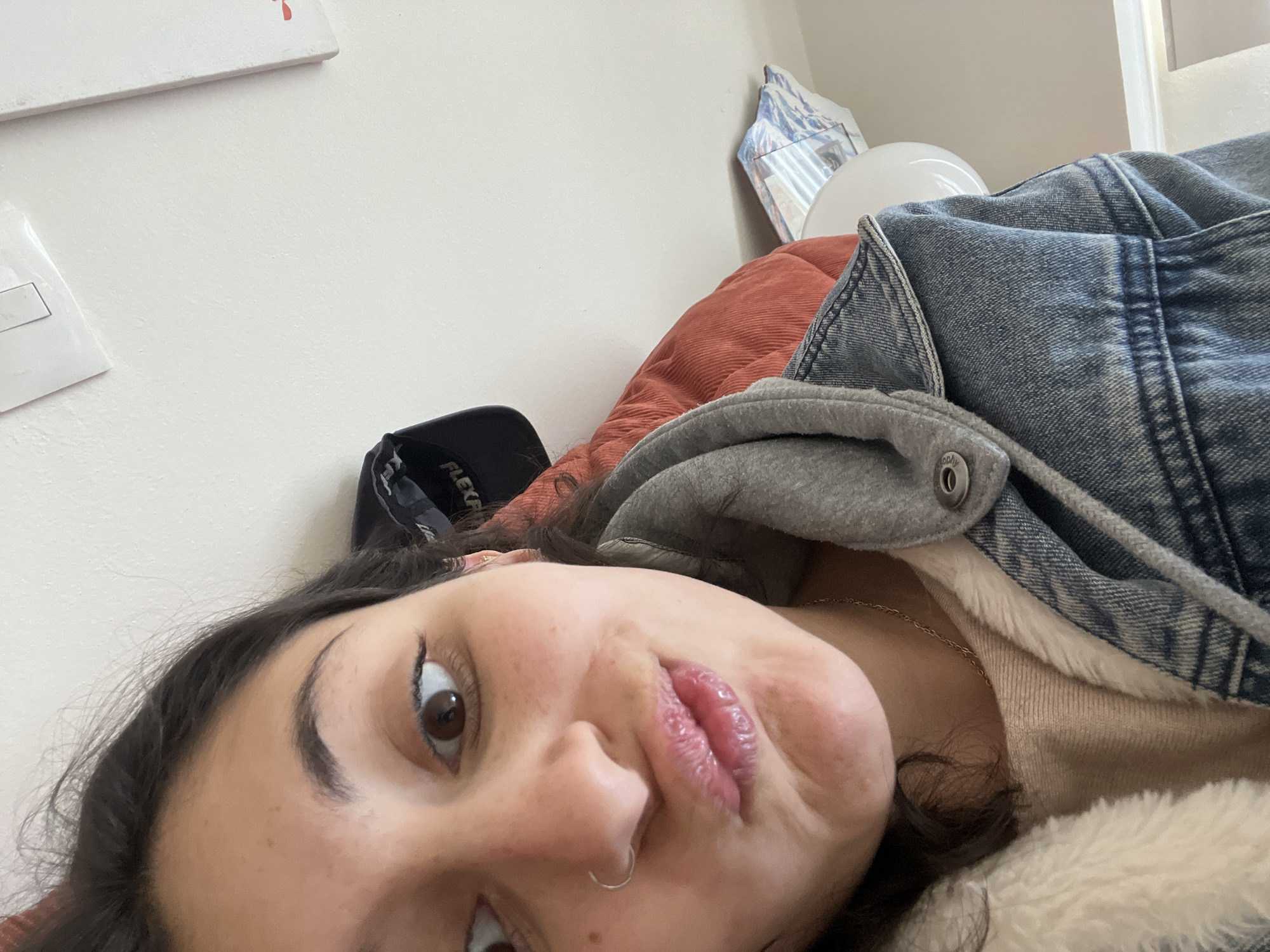 A woman with dark hair is lying down, taking a selfie. She is wearing a denim jacket with a gray hooded sweatshirt underneath, and a beige, fuzzy-lined garment beneath that. A black baseball cap sits beside her head. A rust-colored pillow is behind her, and a white object, possibly a lamp, is behind the pillow. The background includes a white wall with an electrical outlet and a framed picture.
There is no information present that could be used to establish a connection to Huixquilucan de Degollado, Mexico.