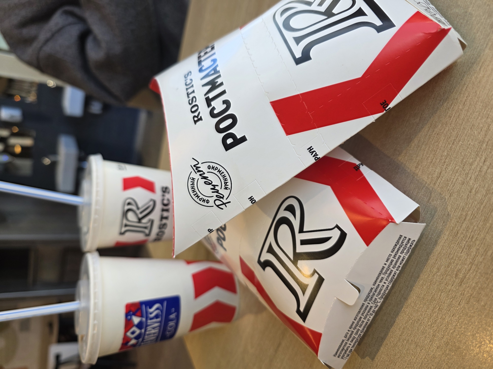 The image displays a fast-food meal from Rostic's, likely in Izhevsk, Russia, set on a light-colored wooden table. Two white cardboard containers, presumably for food, are stacked diagonally across the frame. The top container prominently features the brand "ROSTIC'S" and "РОСТМАСТЕР" (Rostmaster) in black Cyrillic text, alongside "ФИРМЕННЫЙ Рецепт" (Proprietary Recipe) and a large stylized "R" logo. Red chevron-like stripes are part of the design on both containers.
Adjacent to the containers are two white drink cups with straws. The top cup shows the "ROSTIC'S" brand and the "R" logo, while the bottom cup is branded "NERVESS COLA" with the same "R" logo. A blurred dark-colored sleeve of a person is visible in the upper left background.
Small Russian text on the bottom container provides product details: "Наименование, дата и время изготовления продукции, наименование и место нахождения изготовителя пищевой продукции указаны в прилагаемом чеке. Продукцию рекомендуется употребить в пищу сразу после получения. Срок годности и условия хранения продукции +1 час при температуре хранения от +10 до +40°С. Продукция содержит или может содержать" (Name, date and time of product manufacture, name and location of the food product manufacturer are indicated on the attached receipt. It is recommended to consume the product immediately after receiving. Shelf life and storage conditions of the product: +1 hour at a storage temperature of +10 to +40°C. The product contains or may contain...). Other visible partial texts include "РАУН", "OH", and "30E". The scene captures an indoor fast-food dining experience.