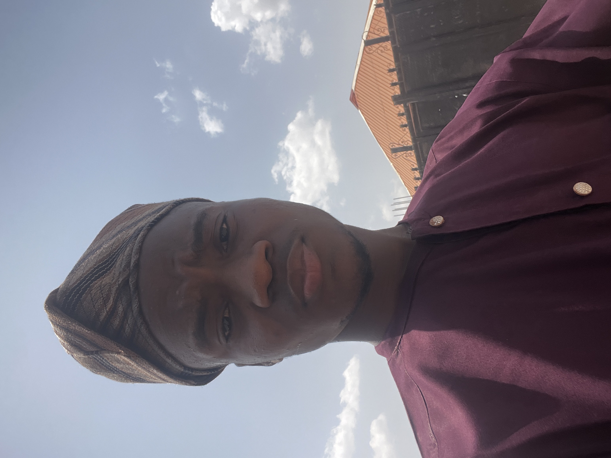 A medium close-up, portrait-style image, originally rotated 90 degrees counter-clockwise, depicts a young man outdoors. The man, with a dark complexion, gazes directly at the camera with a neutral to slightly serious expression. His face appears somewhat shiny, possibly from sweat or natural oils. He wears a patterned, brown traditional cap (likely a Kufi or similar headwear) and a dark red or maroon long-sleeved shirt with two prominent, round, decorative buttons visible on the placket. He has subtle stubble or a short beard on his chin.
The background primarily features a bright, light blue sky with scattered white, puffy clouds. In the upper right portion of the frame, behind the man's right shoulder, a section of a building's roof is visible. The roof features orange or terracotta-colored tiles and some dark, decorative architectural elements, possibly wrought iron. The overall scene suggests a sunny daytime setting with clear skies. The attire worn by the subject, combined with the provided location context, suggests the image was taken in Minna, Nigeria. No visible text is present in the image.