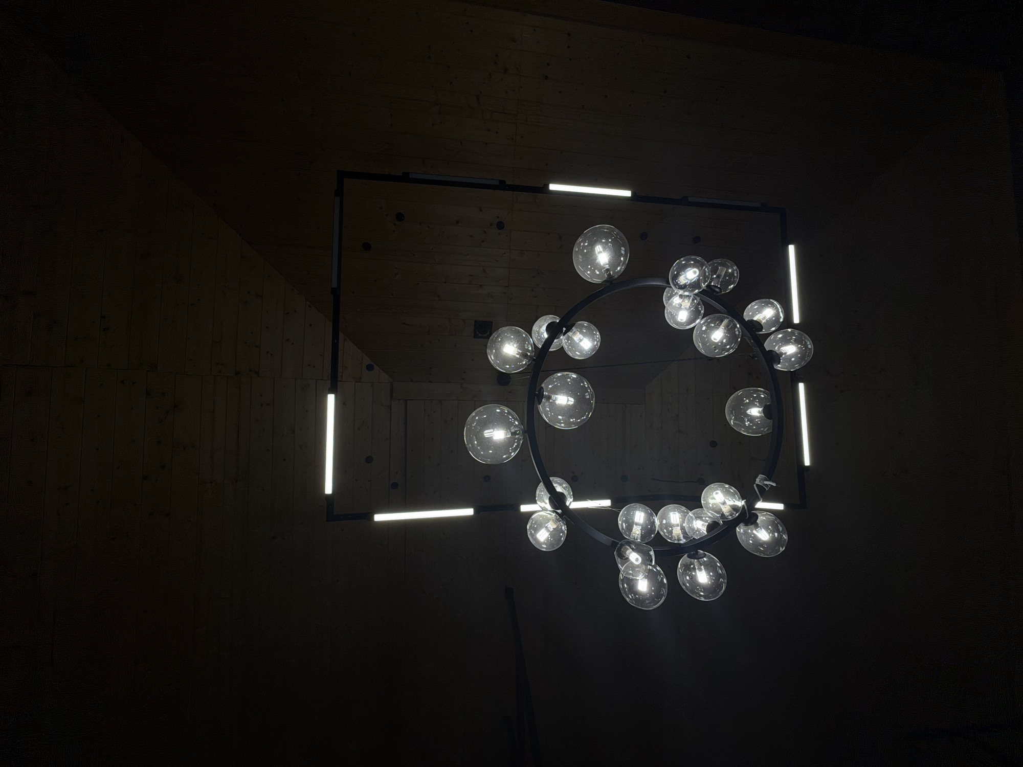 The image depicts a modern chandelier hanging in a dimly lit space. The chandelier has a circular, black metal frame with multiple illuminated glass globes attached around its circumference. A rectangular black metal frame surrounds the circular element, and integrated into this frame are four linear LED light bars. The walls appear to be wood paneled, suggesting an interior space. The low lighting levels and absence of other identifying features make it difficult to determine the exact location, but the prompt indicates it is in Kamennoye, Russia. There are no people visible in the image.