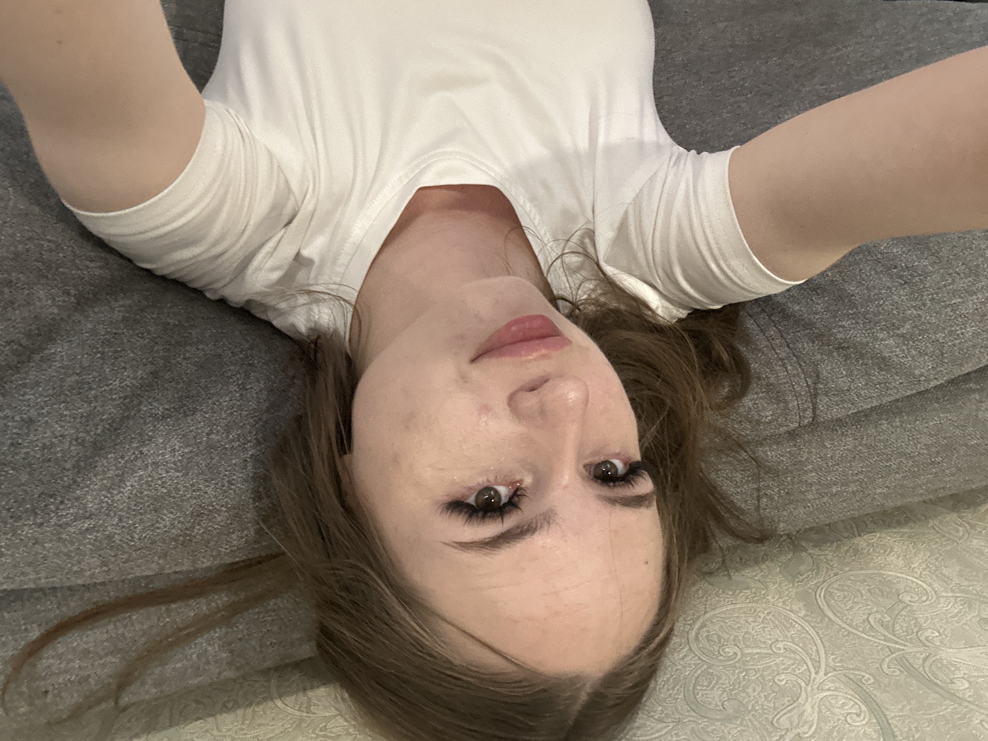 In a close-up, overhead shot, a young woman with long brown hair and dark brown eyes lies on a gray textured surface, possibly a couch or bed. She is wearing a white t-shirt. Her head is tilted, and her gaze is directed upwards, toward the camera. The lighting in the scene is soft and natural, with no harsh shadows. The background is out of focus, but appears to be a continuation of the textured surface. The image is oriented vertically.