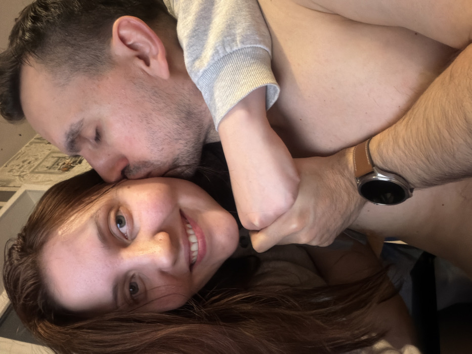 A close-up, intimate photograph captures a couple in a tender embrace, likely taken indoors. The image is oriented at an angle, showing the subjects tilted.
In the foreground, a woman lies down, looking directly at the camera with a bright, open-mouthed smile. She has long, flowing brown hair that cascades across the bottom of the frame, and light-colored eyes. Her skin appears fair.
Behind and slightly above the woman, a man is seen with his eyes closed, gently kissing her forehead or hair. He has short, dark hair and noticeable stubble on his face. He is wearing a light grey long-sleeved garment, possibly a sweatshirt, over his upper body, with a portion of his bare torso visible.
Their arms are intertwined in a hug, conveying affection. The woman's arm, partially visible with a light-colored sleeve, is wrapped around the man's arm, with her hand gripping his forearm. The man's forearm, which is visibly hairy, is also seen, adorned with a smartwatch featuring a round silver-colored casing and a light brown leather strap on his left wrist. The watch face appears dark.
The background is softly blurred, revealing a light-colored surface with a subtle, indistinct pattern, possibly wallpaper or a fabric headboard. The overall atmosphere of the image is one of warmth, happiness, and intimate connection between the two individuals. No text is visible in the image.
