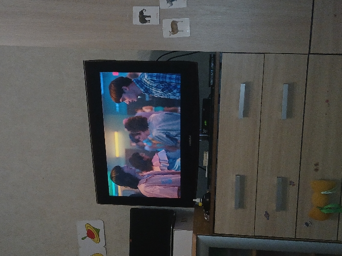 A television displaying a scene with multiple people is mounted on a wall beside a wooden cabinet with silver handles. Playing cards with animal illustrations are placed on the wall near the TV. A black box, likely a DVD player, is positioned underneath the TV. More playing cards are near the bottom of the screen, and a stuffed animal hangs off the side of the cabinet.