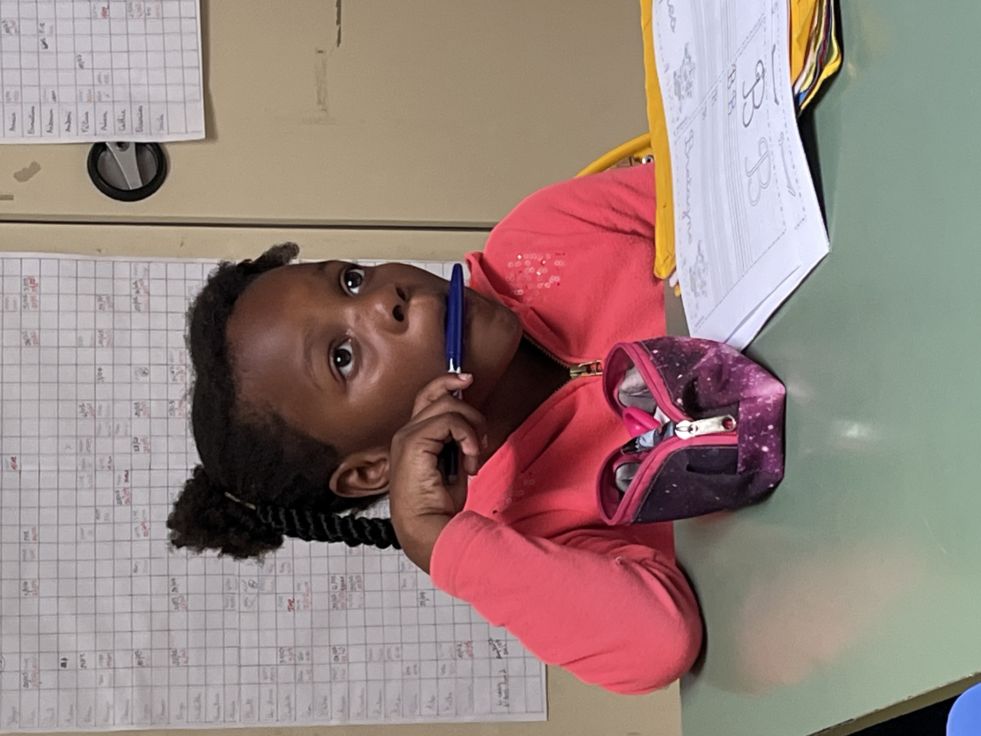 A young Black girl is seated at a light green table, deeply focused on an activity in what appears to be a classroom in La Fouillouse, France. She is wearing a coral-colored long-sleeved top with sparkling embellishments on the shoulder, and her dark hair is styled in braids. Holding a blue pen near her mouth, she gazes upwards with a thoughtful and intent expression, her eyes wide. On the table directly in front of her is a sheet of paper displaying handwritten letters, prominently featuring two large "B"s and the word "Bretagne" (Brittany). To her right, an open purple and white galaxy-print pencil case, containing various school supplies like scissors, rests on the table. The background consists of light-colored walls or cabinets adorned with several grid-patterned charts, possibly classroom schedules, attendance sheets, or planning boards. These charts contain numerous handwritten names such as "Amelie," "Anthony," "Vilma," "Celine," "Damien," and "Remi," along with various dates and other largely illegible notes. The image captures a moment of quiet concentration during a learning or school-related task.