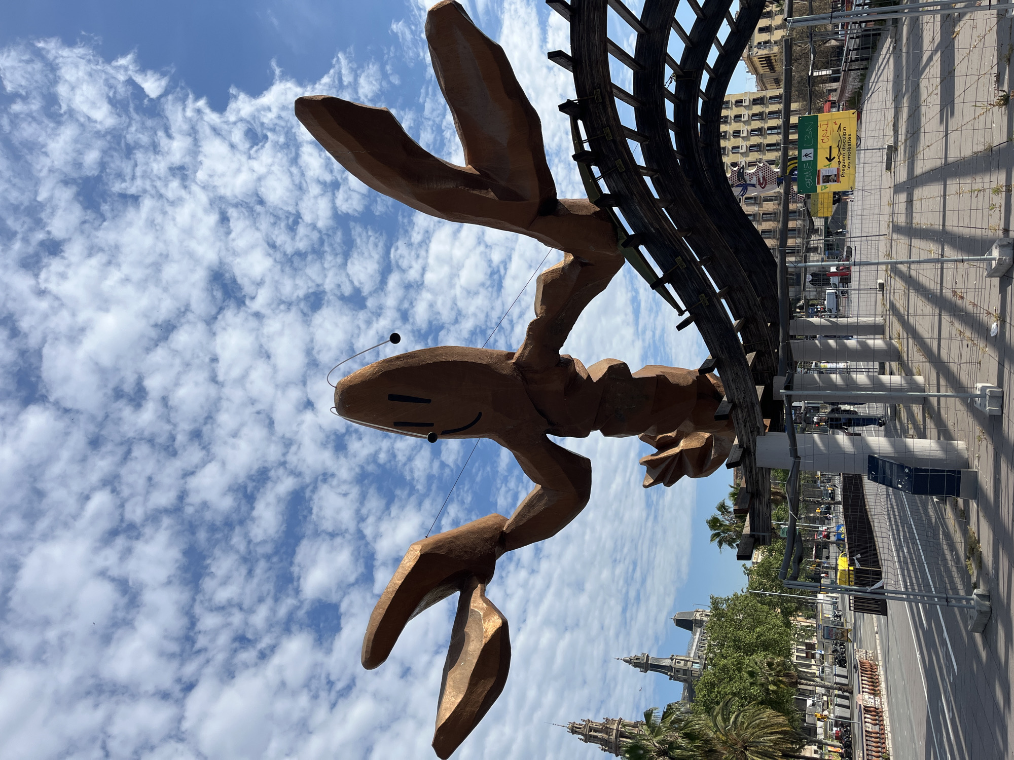 The media file depicts a vibrant urban scene, presented rotated 90 degrees clockwise from its intended upright orientation. The central focus is a large, whimsical sculpture of a lobster, appearing to be crafted from a bronze or copper-toned material, set against a bright blue sky filled with scattered white cumulus clouds. The lobster features large claws, antennae, and a stylized, smiling face with distinct eyes. Adjacent to the lobster's back is a curved wooden lattice structure, possibly part of the same artistic installation or nearby architecture.
The background reveals a bustling cityscape typical of Barcelona, Spain. Multi-story buildings with classic European architectural details line a street where cars and motorcycles are visible. Lush green trees, including characteristic palm trees, dot the urban landscape. Prominent among the buildings is one with ornate spires, characteristic of historic Barcelona architecture.
In the foreground and midground, temporary metal construction fencing separates the viewer's perspective from the street. Behind this fencing, a yellow rectangular sign is visible. It contains black text and arrows, with decipherable phrases including "Obres C.B.I." and "Prega d'ampliar les molèsties," which is Catalan for "Please excuse the inconvenience," indicating ongoing construction or maintenance. A smaller sign with blue and green text is partially visible behind the yellow one.
The scene is illuminated by bright natural daylight, suggesting it is daytime under clear and sunny weather conditions. The overall impression is that of a lively city featuring a distinctive and iconic piece of public art, known as the Gambrinus lobster sculpture, set within an active urban environment undergoing some form of work or event setup due to the fencing.