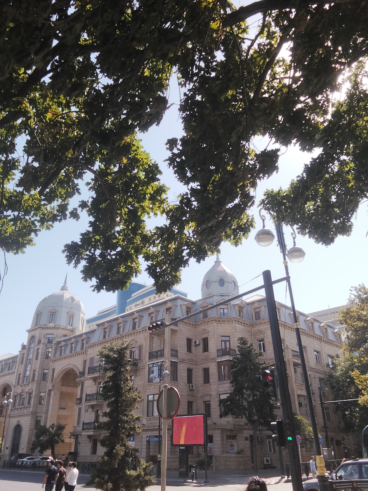 The image captures a street scene in Baku, Azerbaijan, looking up towards a grand, multi-story building with classical architectural elements, including arches, ornate cornices, and mansard roofs. A prominent dome with a spire tops the left section of the building, suggesting historical significance or a landmark status. A modern blue glass building with "Hilton" signage is visible behind it, indicating a blend of old and new architecture in the city. Overhead, large tree branches with lush green leaves frame the upper portion of the image, with sunlight filtering through, creating a dappled effect against the clear blue sky. In the foreground, a street lamp with two globe lights stands tall, and a traffic light shows a red signal. Several people are walking on the sidewalk and a few cars are parked along the street. A tall, slender evergreen tree stands in front of the building. The overall scene depicts a sunny day in an urban environment with significant architectural features.