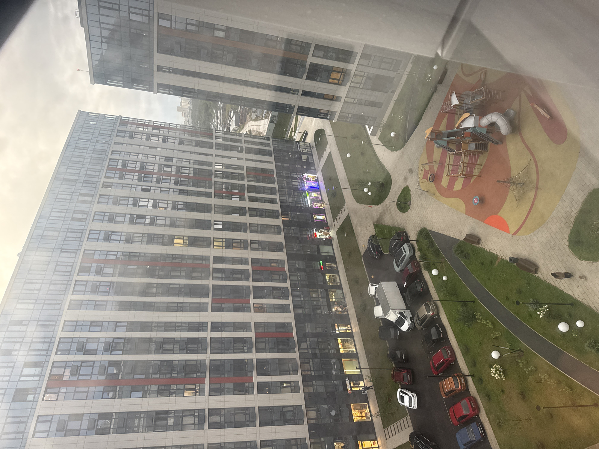 The image captures an aerial view of a modern residential complex in Energetik SNT, Russia, on a cloudy day. Two tall, contemporary apartment buildings with extensive glass facades dominate the scene, featuring a pattern of light-colored panels and red vertical accents. The buildings are situated adjacent to each other, with one angled slightly towards the other.
In the foreground and middle ground, a residential courtyard is visible. A children's playground with colorful rubberized surfacing and various play structures, including a slide, swings, and climbing frames, is a prominent feature. Paved walkways wind through grassy areas with scattered trees and decorative spherical lights. Several parked cars of various colors and a white delivery truck are visible on a paved surface adjacent to one of the buildings. A few individuals are present in the scene: one person walks a dog on a pathway, and other figures are seen near the ground-floor commercial spaces of the buildings. The ground floor of the buildings appears to house retail establishments, indicated by illuminated storefronts and signage. The overall environment suggests a newly developed urban residential area.