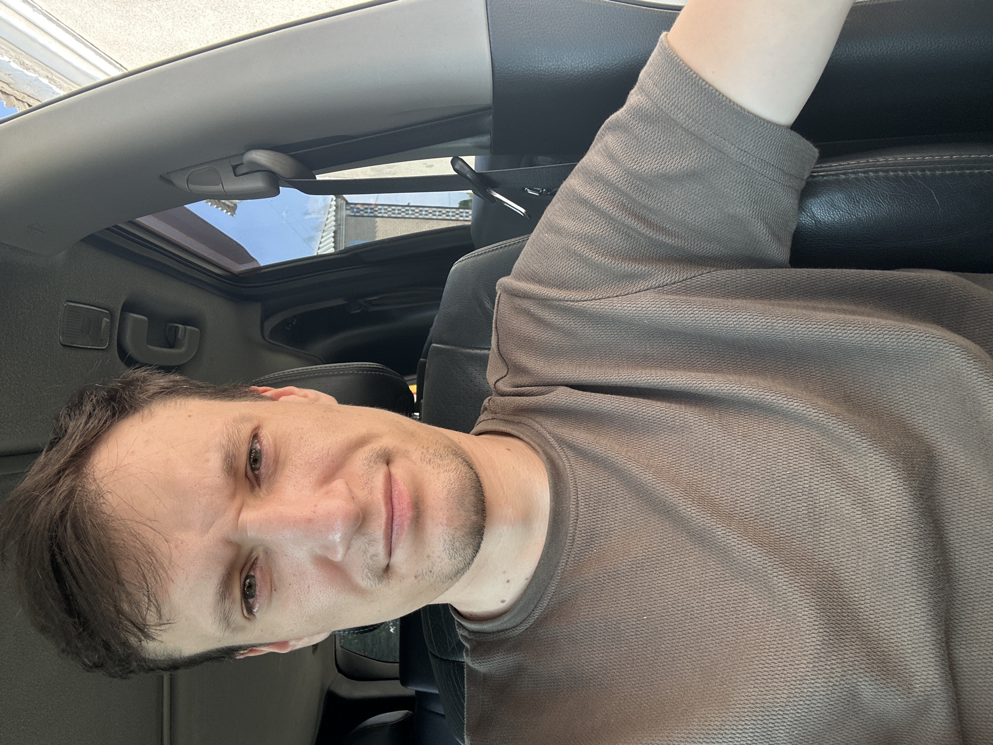 A man is sitting in the driver's seat of a car, with a selfie-style shot. He has short, dark brown hair and is wearing a brown t-shirt. The interior of the car is visible, including black leather seats and a gray interior panel. Outside the car window, a building and trees can be seen. The scene appears to be outdoors on a sunny day. The image was taken in the city of Shatava, Ukraine.