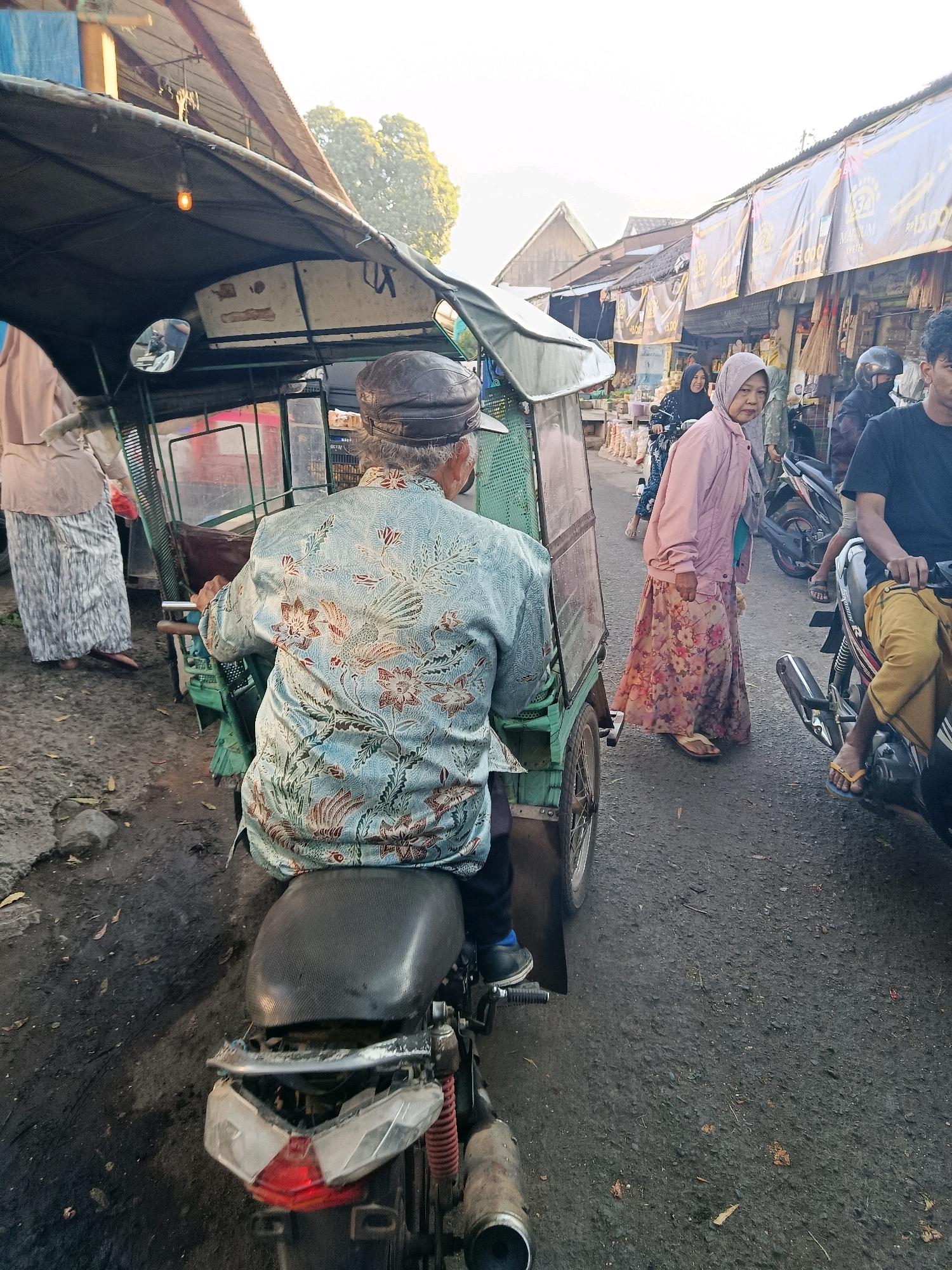 The image depicts a street scene in Bululawang, Indonesia, likely a marketplace. In the foreground, an elderly man wearing a patterned blue shirt and a dark cap is seated on a motorcycle, facing away from the camera. He is operating a three-wheeled vehicle attached to the motorcycle, with a green frame and a dark canopy. To the right of the man, a woman wearing a pink jacket and a floral skirt walks past, looking towards the camera. Behind her, other people are visible on motorcycles, and stalls line the street, indicated by banners and goods. The ground is paved and appears somewhat dirty. The lighting suggests it is daytime, possibly late afternoon due to the warm tones in the sky. The overall atmosphere appears to be one of a busy local commercial area.