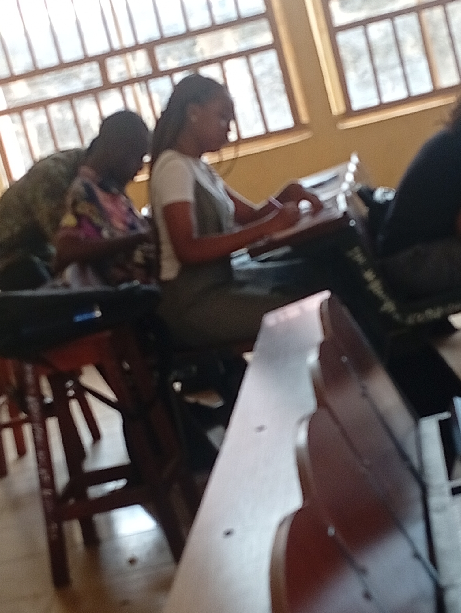 The image depicts a classroom setting in Ukukwa, Nigeria, with two individuals seated at desks. A young woman in a white shirt and green overalls is actively writing or drawing on a surface in front of her. To her left, another individual, partially obscured, appears to be wearing a patterned shirt and is also engaged in an activity at a desk. The environment suggests a school or educational institution, with windows visible in the background, allowing natural light to illuminate the scene. The presence of desks and seating arrangements further reinforces this context.
