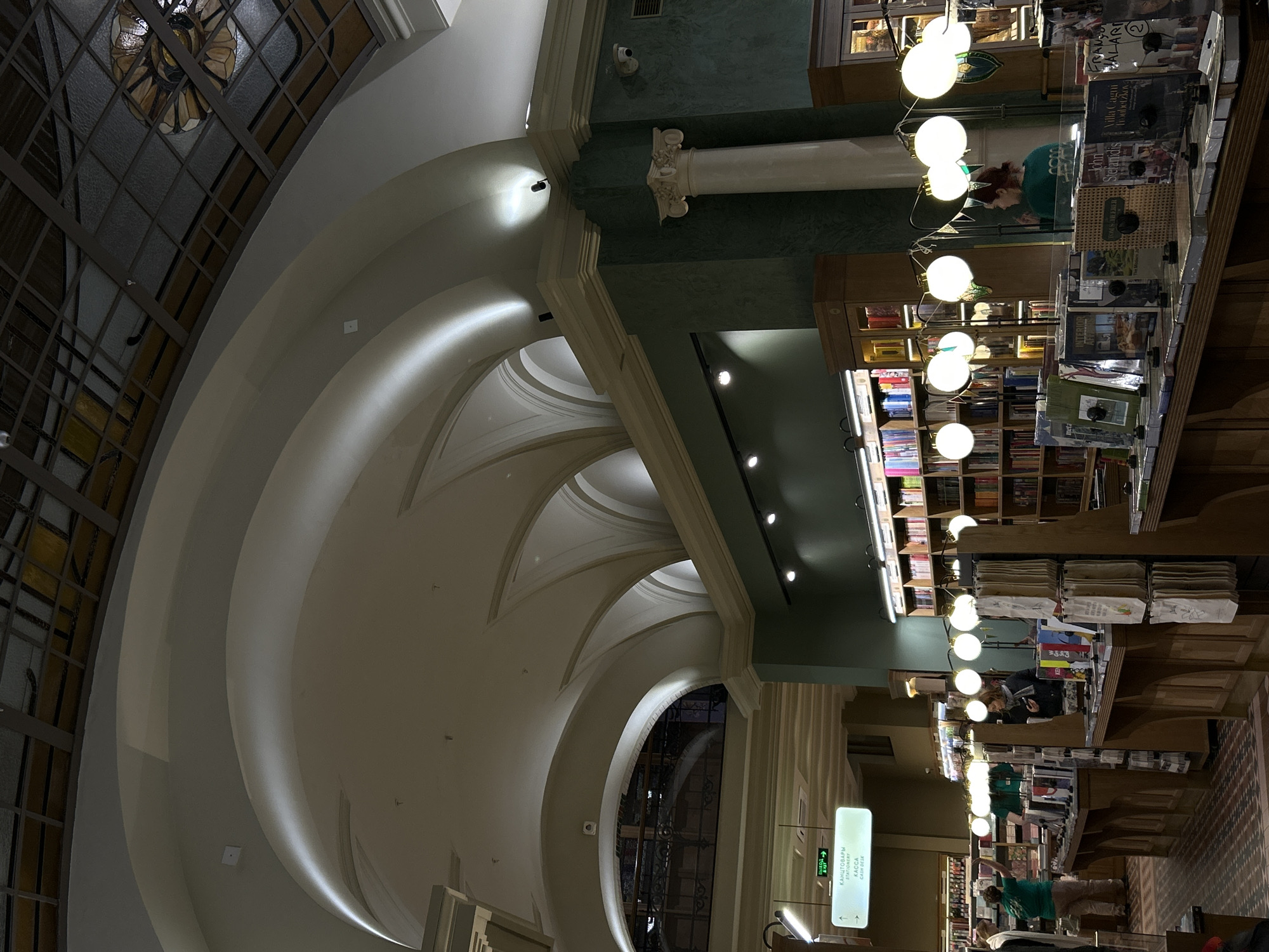 This image captures the interior of a grand bookstore or library in Saint Petersburg, Russia, characterized by its elegant architecture and warm lighting. The perspective looks across a spacious hall, dominated by high, vaulted white ceilings with decorative cornices and soft, indirect lighting.
On the left, a large, ornate arched window with a grid pattern allows natural light into the space. A classical white column with a decorative capital stands on the right, set against a dark teal or green textured wall. The main area is filled with numerous wooden bookshelves and display tables laden with books. Overhead, rows of glowing spherical light fixtures illuminate the displays, creating an inviting atmosphere.
Several people are visible throughout the space, primarily engaged in browsing the books. One individual wearing a green top is seen leaning over a display table, seemingly examining items. Other patrons are visible further down the aisle.
Notable details include various books on display, with some titles partially visible, such as "Villa Cagni", "John Stefanidis", and "FRANÇALANI". Near the back, a light-up sign in both Cyrillic and English reads "КАНЦТОВАРЫ / STATIONERY" and "KACCA / CASH DESK," accompanied by an arrow indicating an exit. The ambiance suggests a refined and cozy environment conducive to reading and exploration.