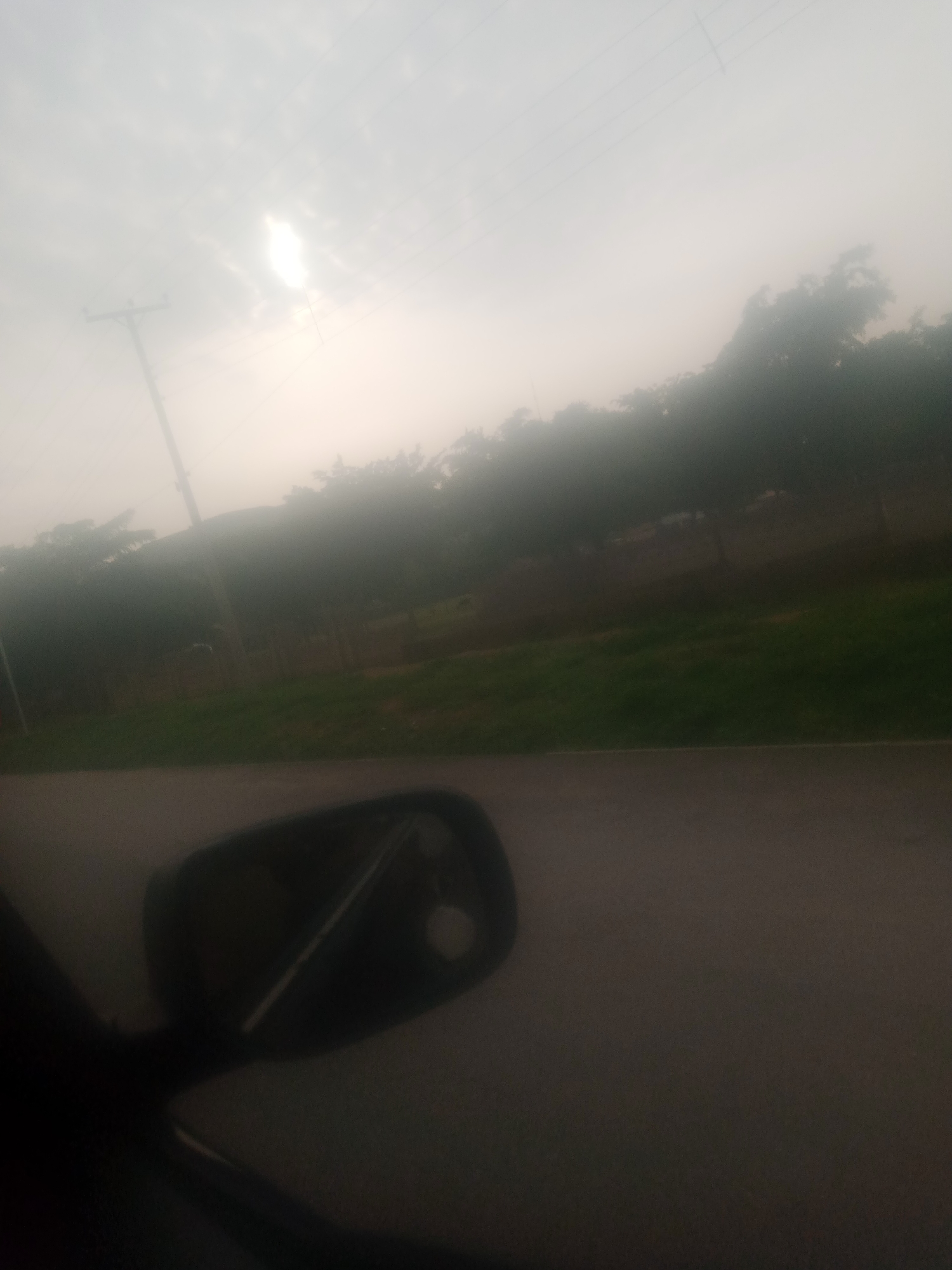 This is a slightly hazy, outdoor, eye-level shot taken from inside a vehicle, likely a car, looking out towards a roadside scene. The prominent foreground element is the dark silhouette of a car's side mirror on the left side of the frame, reflecting a bright, out-of-focus light source, possibly the sun. Beyond the mirror, a gray asphalt road surface is visible.
The midground features a line of trees and vegetation. A single utility pole with wires is visible on the left, standing against a pale, overcast sky. The trees are dense and appear to be lining a roadside embankment. A strip of green grass runs parallel to the road. In the background, the sky is bright and hazy, with a bright patch of light, suggesting the sun is obscured by fog or clouds. The overall impression is a view from a moving vehicle on a somewhat overcast day, possibly during dawn or dusk, in a semi-rural or suburban area. The scene is located in Jalingo, Nigeria.